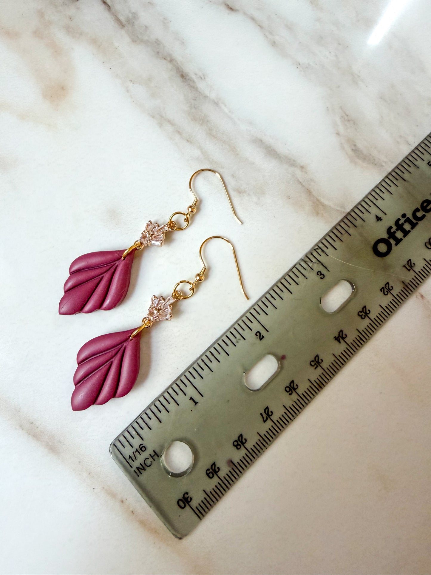 A pair of dangling earrings with magenta leaf-shaped charms and gold hooks, displayed next to a ruler on a light marble surface. 2 1/2 inches in length.