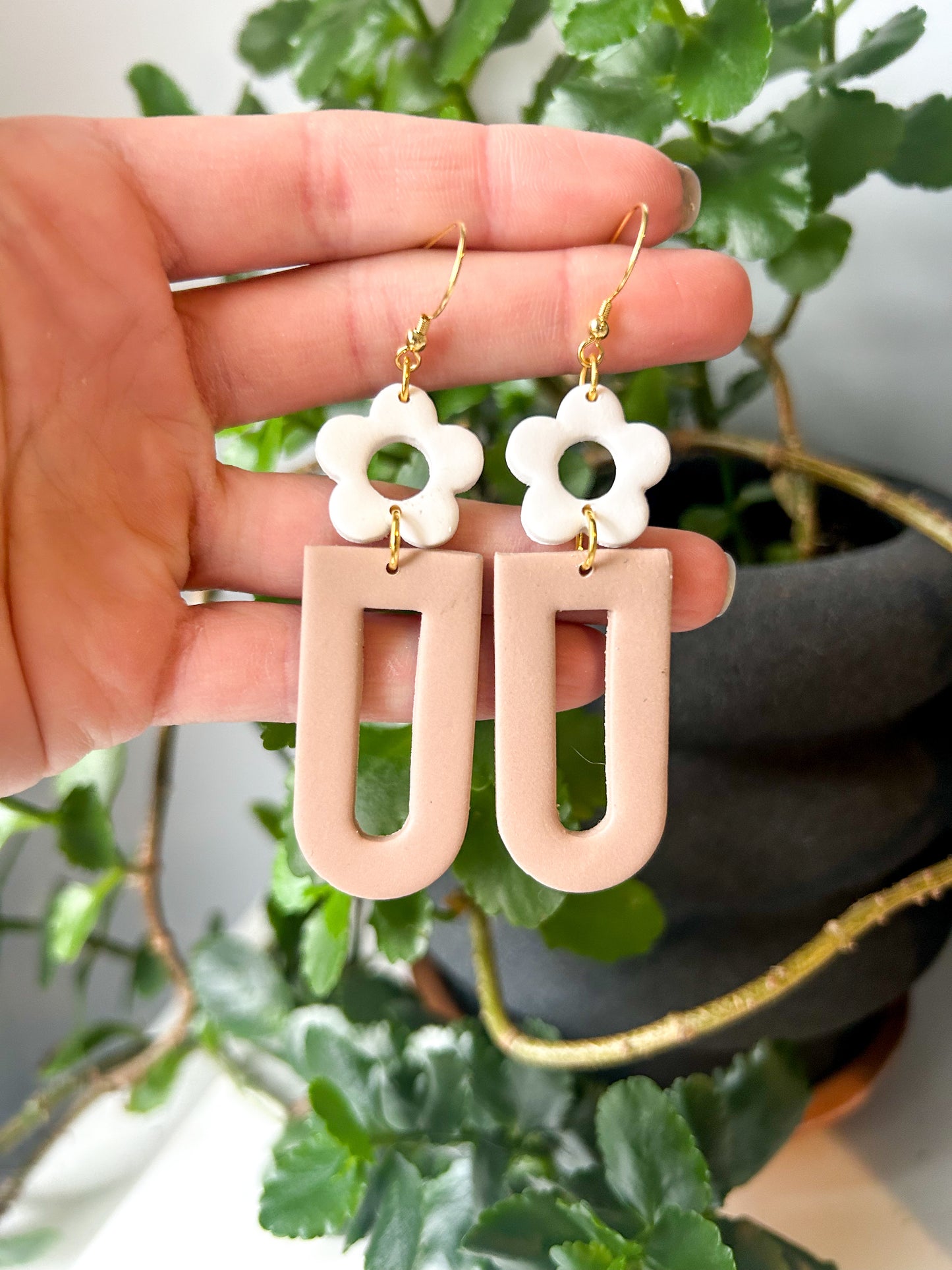 A hand holding a pair of earrings, with the earring on the top featuring a white flower design and the bottom in a pale brown color, suspended from gold-colored hooks.