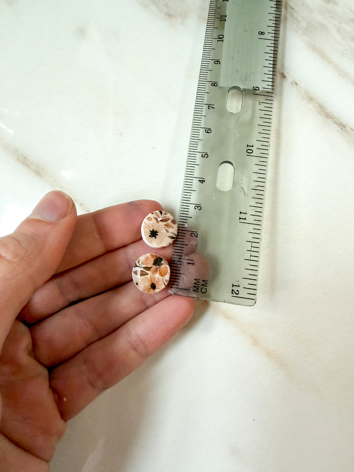 A hand holds a pair of small round stud earrings with a beige and brown floral pattern next to a clear plastic ruler on a light marble surface. About 1cm in length