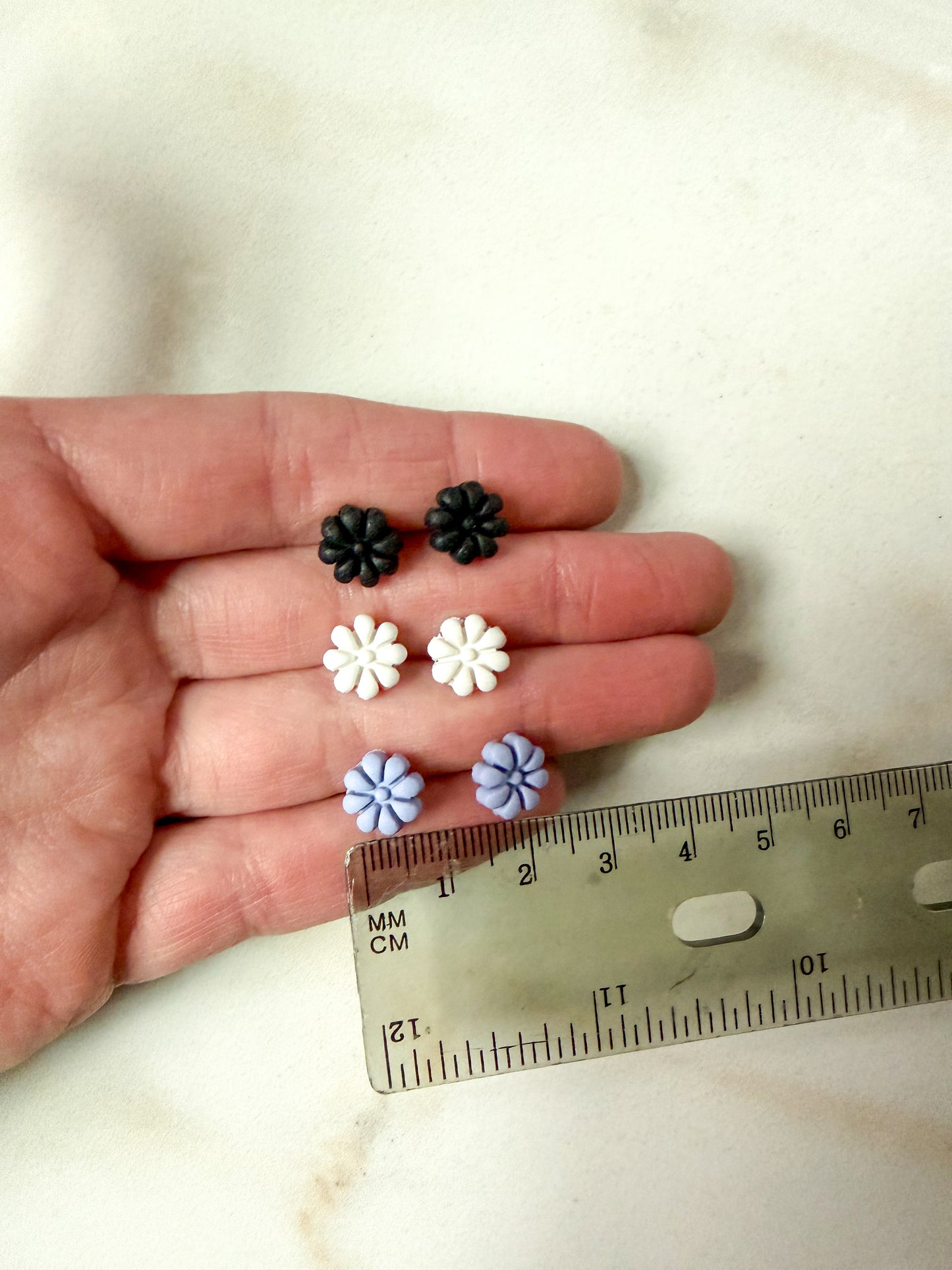 Small black, white, and purple flower-shaped earrings held in a hand with a ruler for scale. About 1cm long.