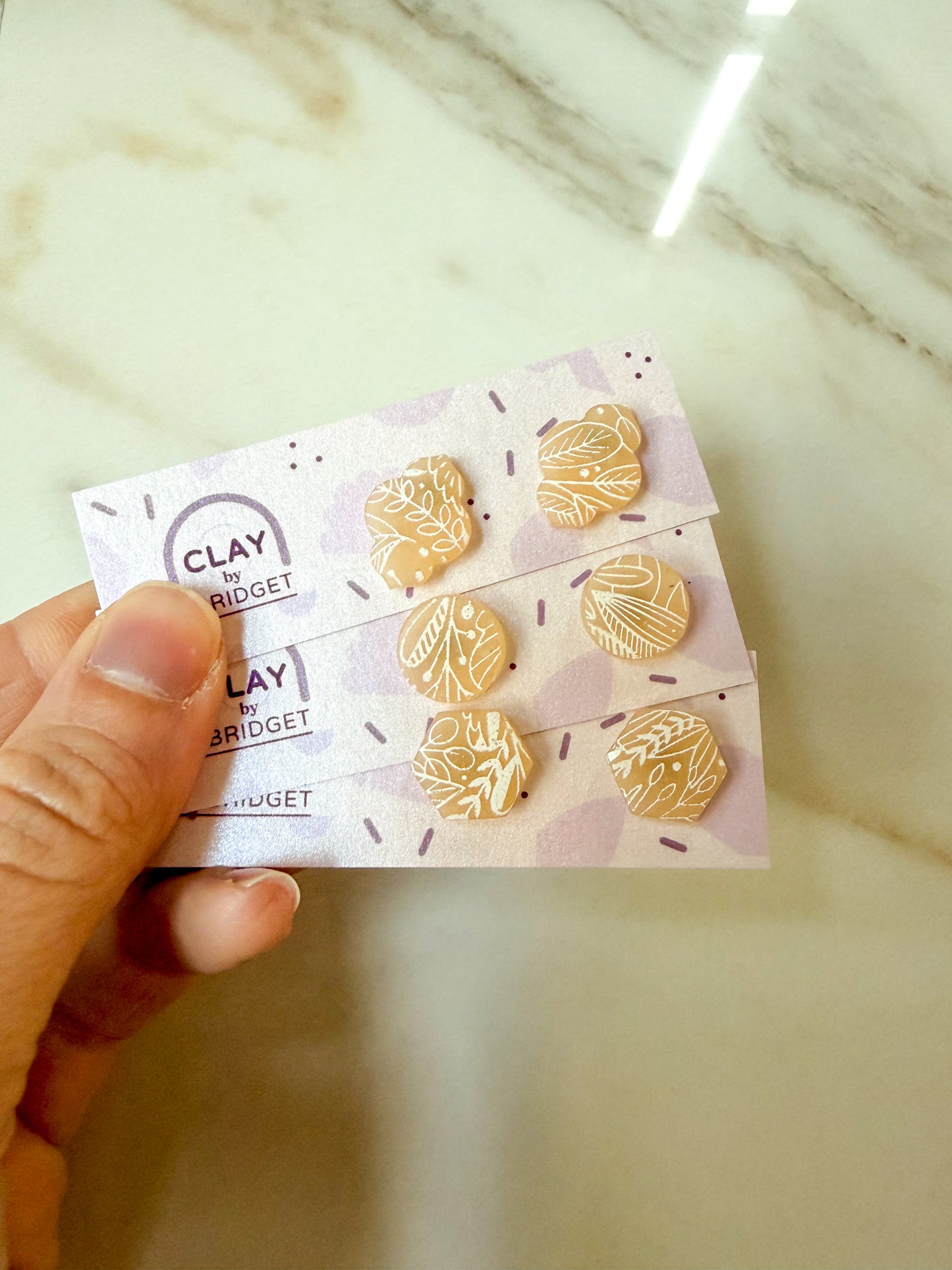 A hand holds a small display card showing three pairs of gold-toned clay earrings with white floral designs, against a light marble background.