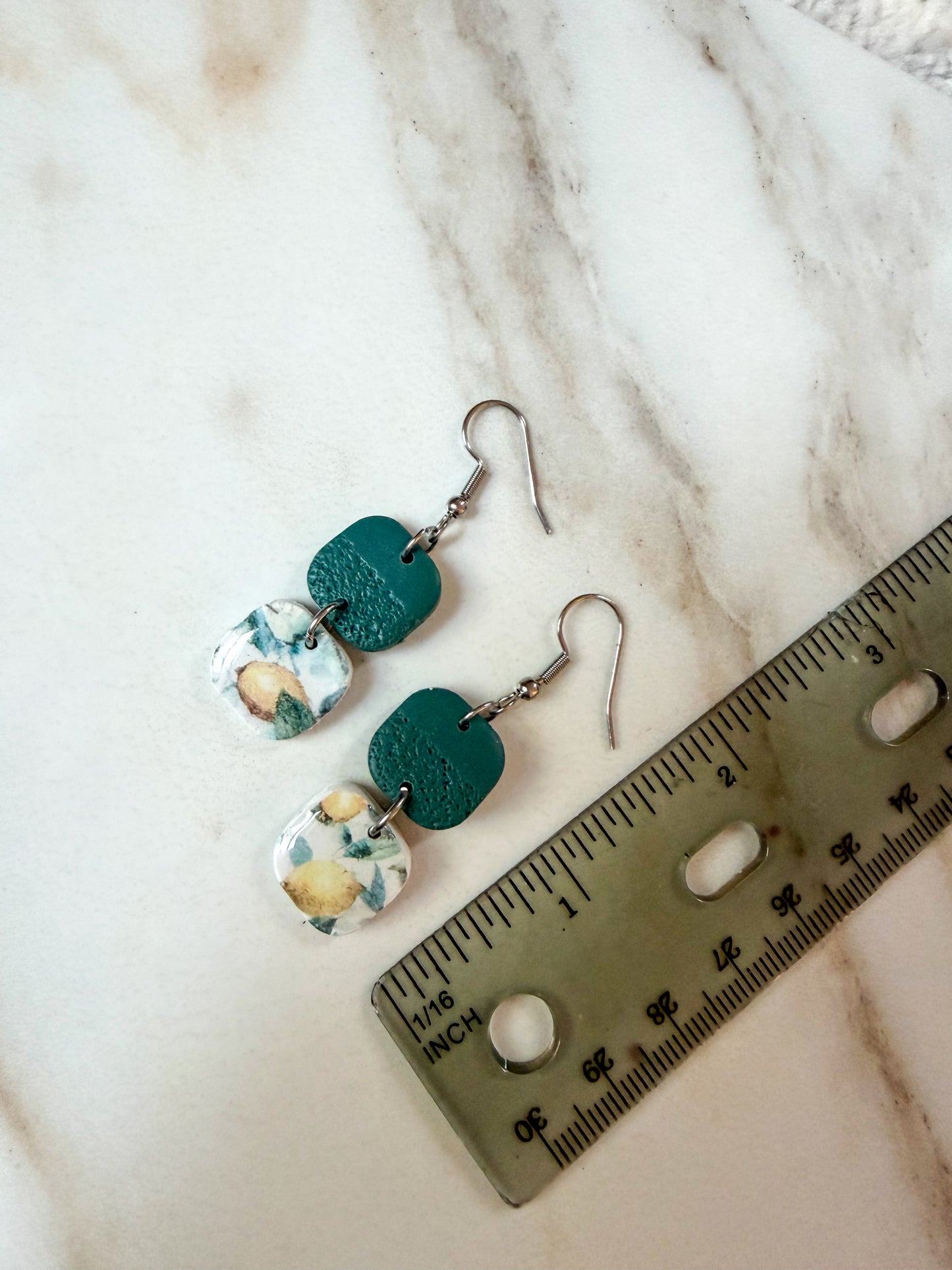 A pair of handmade dangle earrings with textured teal green and white lemon patterned pieces, displayed next to a metal ruler on a light marble surface. About 2 inches in length.