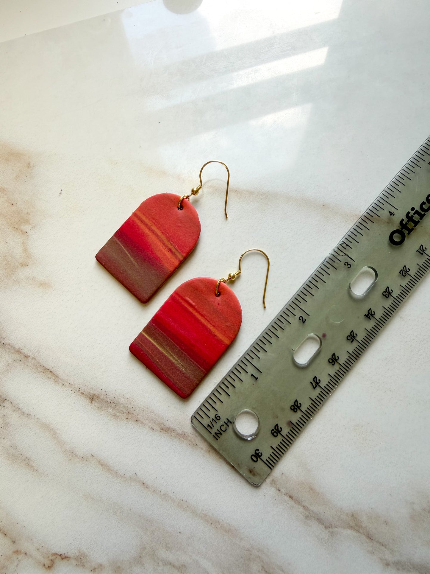 Earrings laying on a marble surface next to a ruler for scale. The design resembles a sunset with colors fading from orange to brown. They are about 2 1/2 inches long.