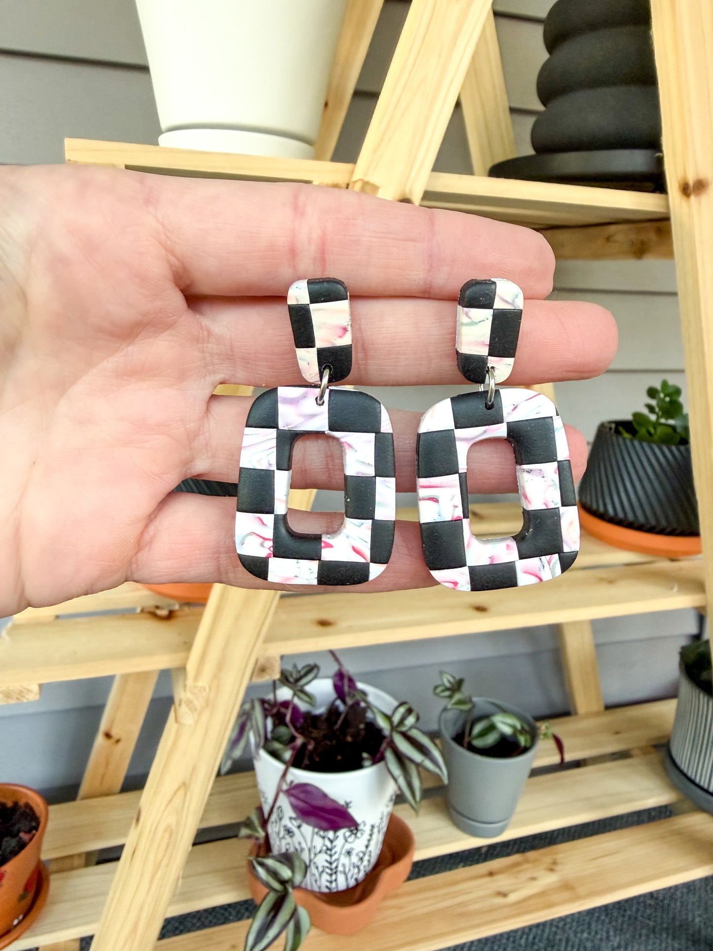 Checkered earrings held by a hand with a wooden shelf and plants in the background