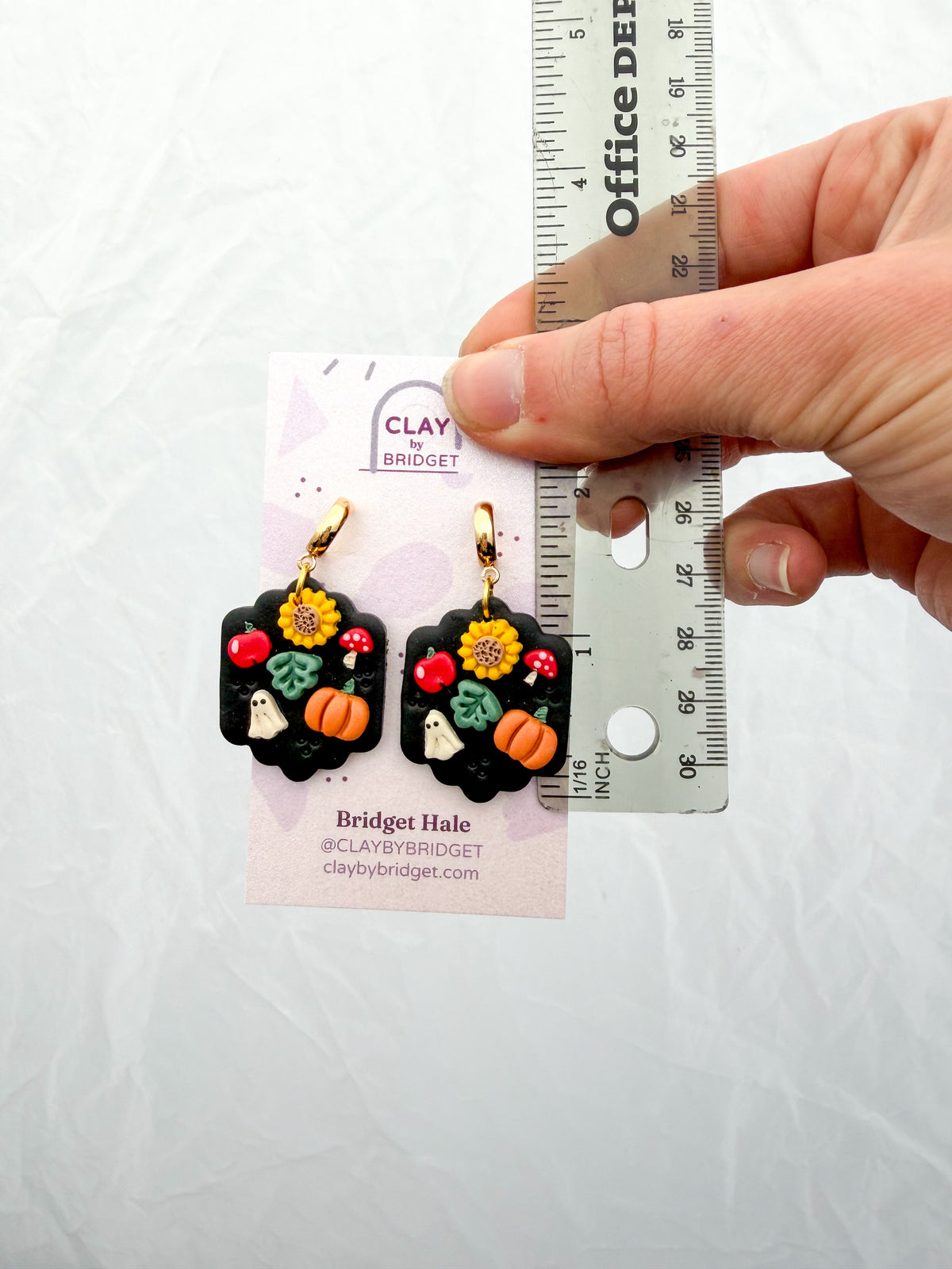 A hand holds a pair of small black polymer clay earrings with autumn garden details, next to a ruler and a branded display card, on a light background. Around 2 inches long.