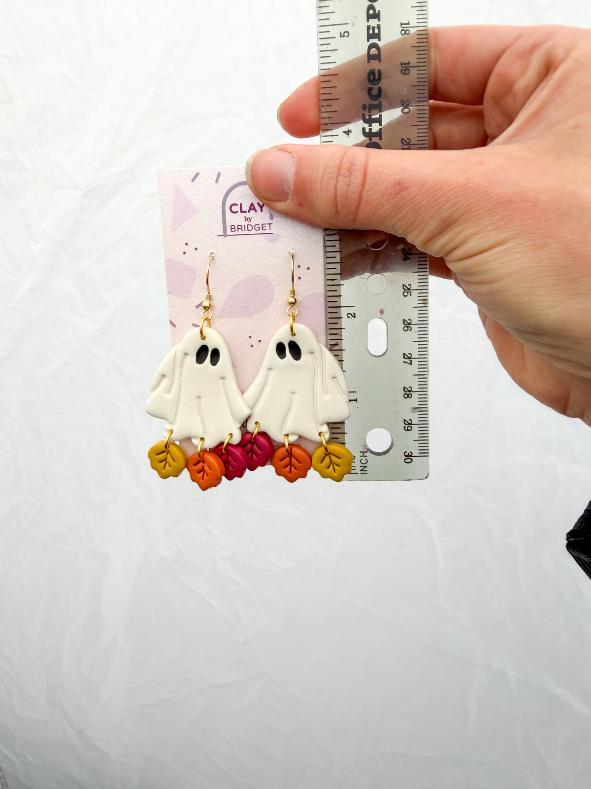 A hand holds a pair of Halloween-themed clay Earrings with white ghost charms and colorful autumn leaves, shown next to a ruler. About 2.5 inches tall.