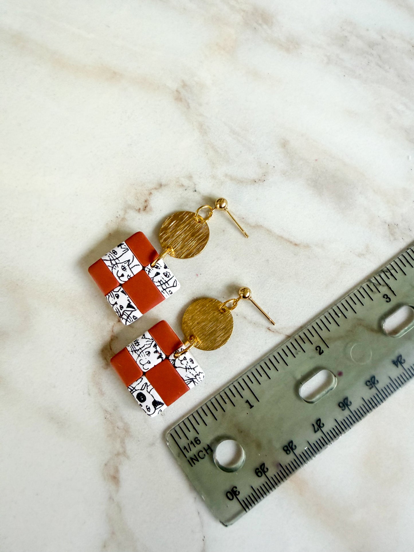 Pair of earrings with terracotta and white patterned stones and gold accents on a marble surface with a ruler for scale.