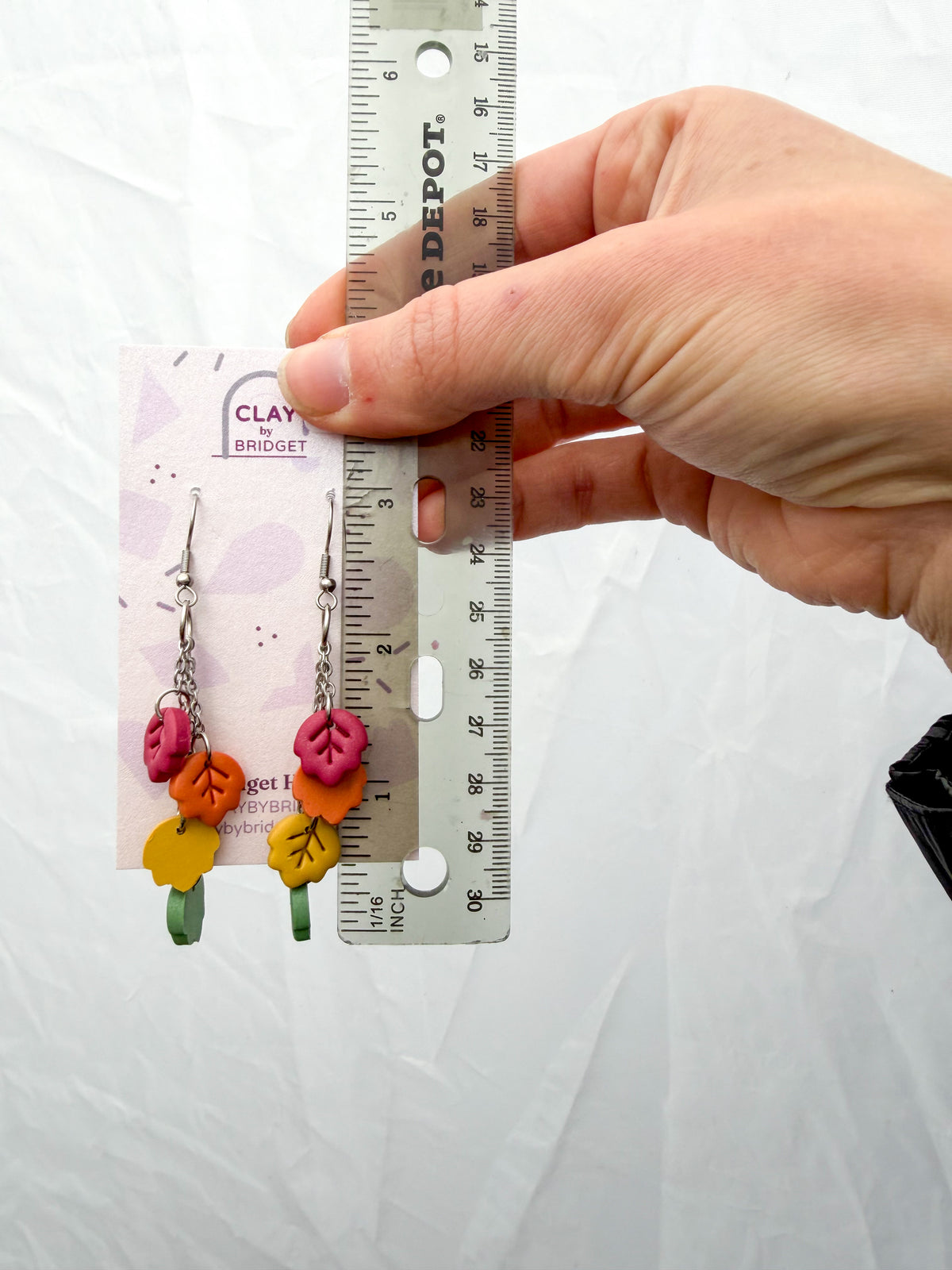 A hand holds a pair of colorful clay leaf dangle earrings on a display card next to a ruler for scale, against a white fabric background. Around 3 inches in length.