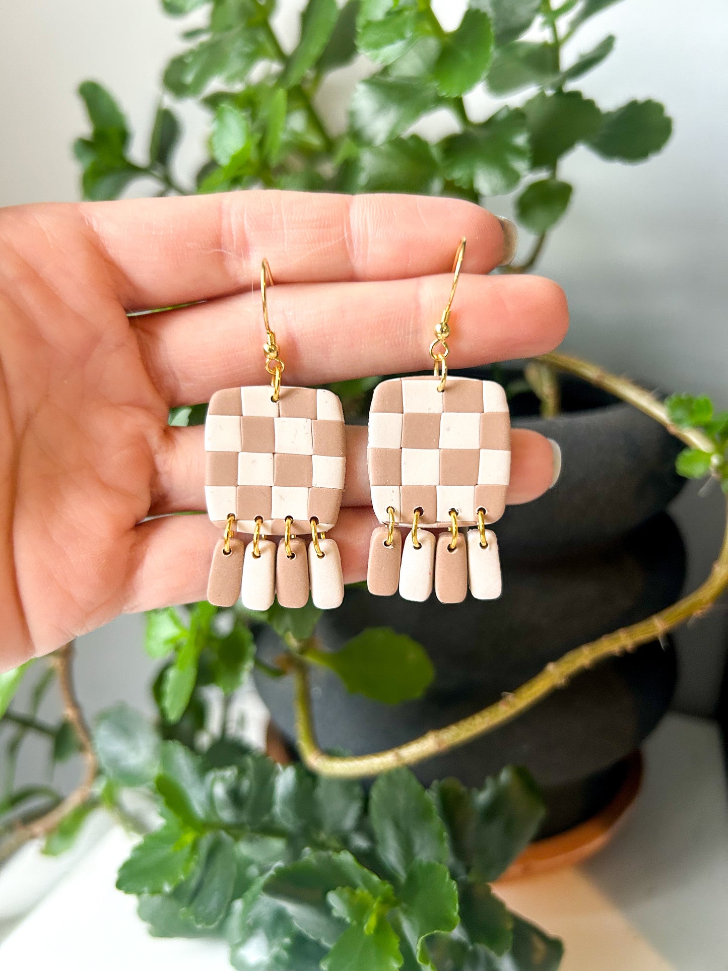 A hand holding a pair of brown and cream checker square fringe dangle earrings with gold-colored hooks, in front of a potted plant.