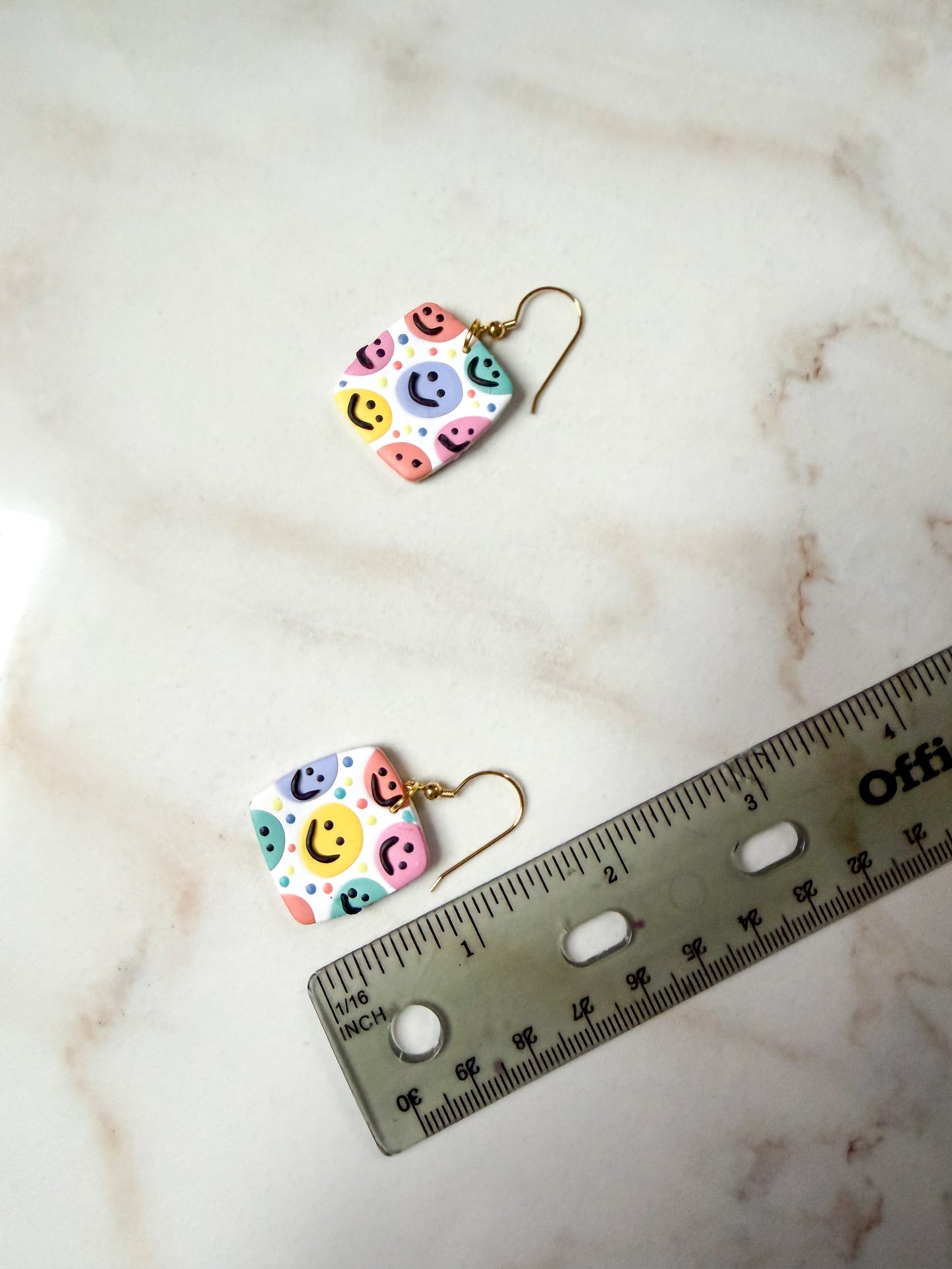 Colorful smiley face earrings on a marble surface with a ruler for scale. A little under 2 inches long.