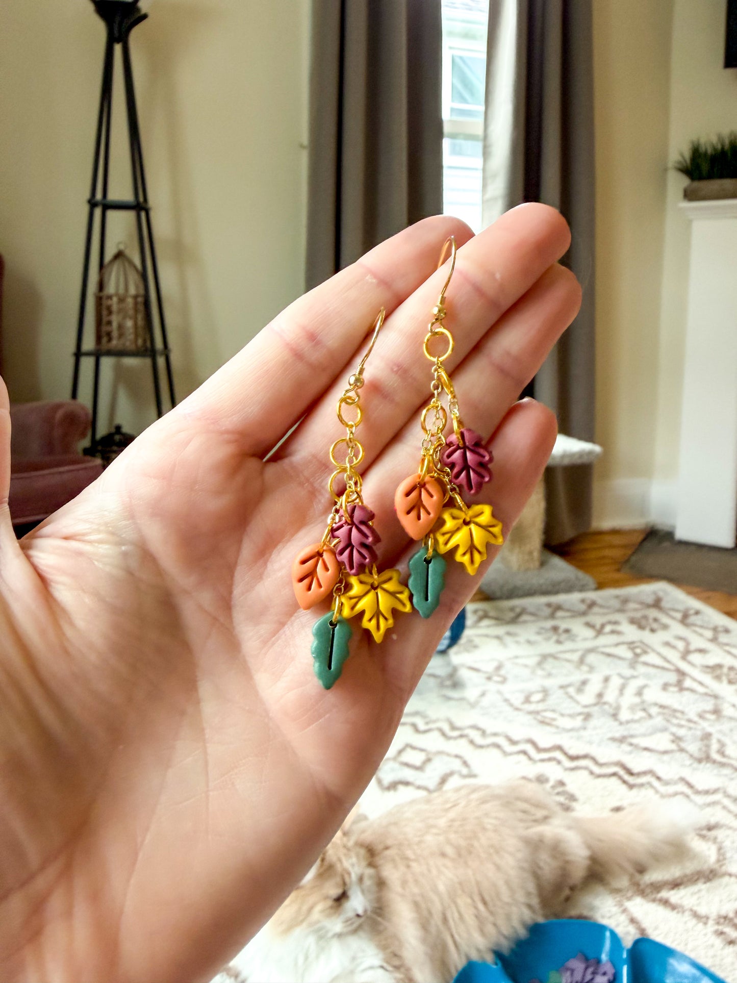 A hand holds a pair of gold-tone dangle earrings with colorful autumn leaf charms in orange, yellow, purple, and green, shown in a home setting with a cat in the background.