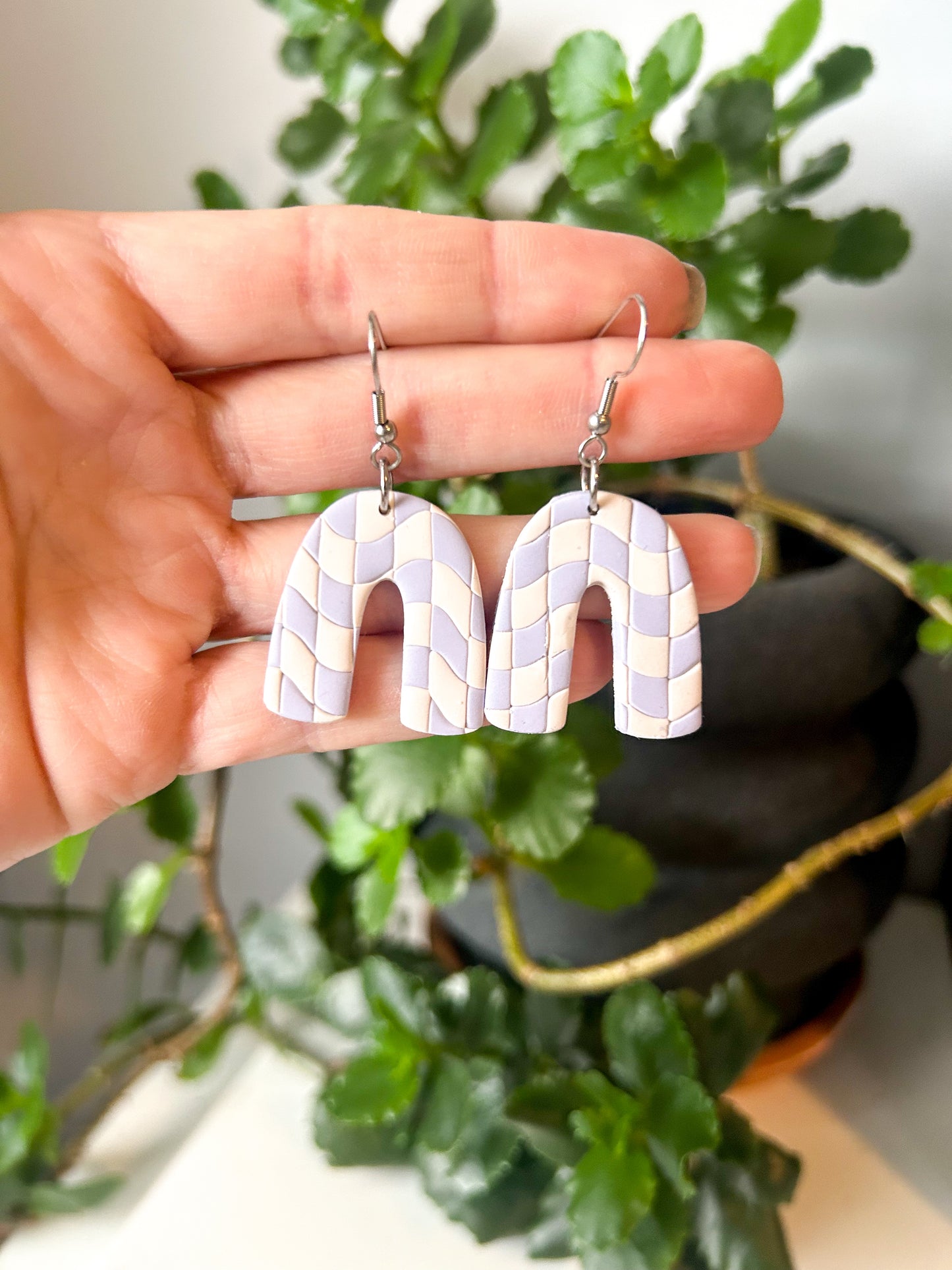 A pair of cream and periwinkle checker patterned arch-shaped earrings held in a hand, with a plant in the background. Swirled checker.