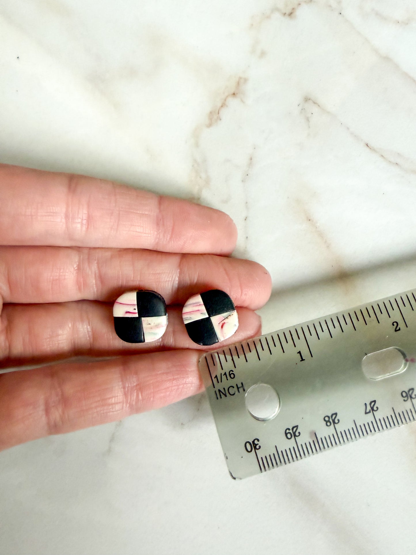 Black and white checkered earrings held between fingers with a ruler for scale on a marble background. About half an inch wide
