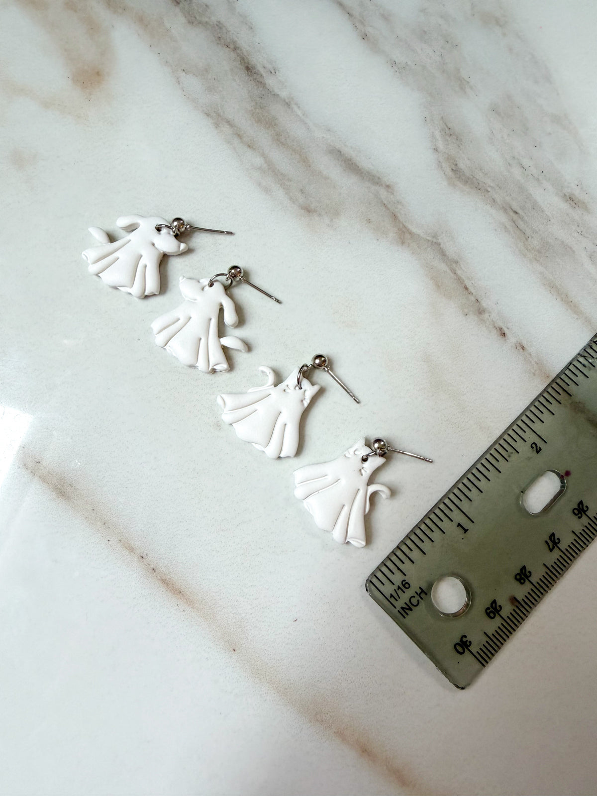 Four small white ghost-shaped earrings with silver posts are arranged on a white marble surface next to a metal ruler for scale. About 1 inch in length.