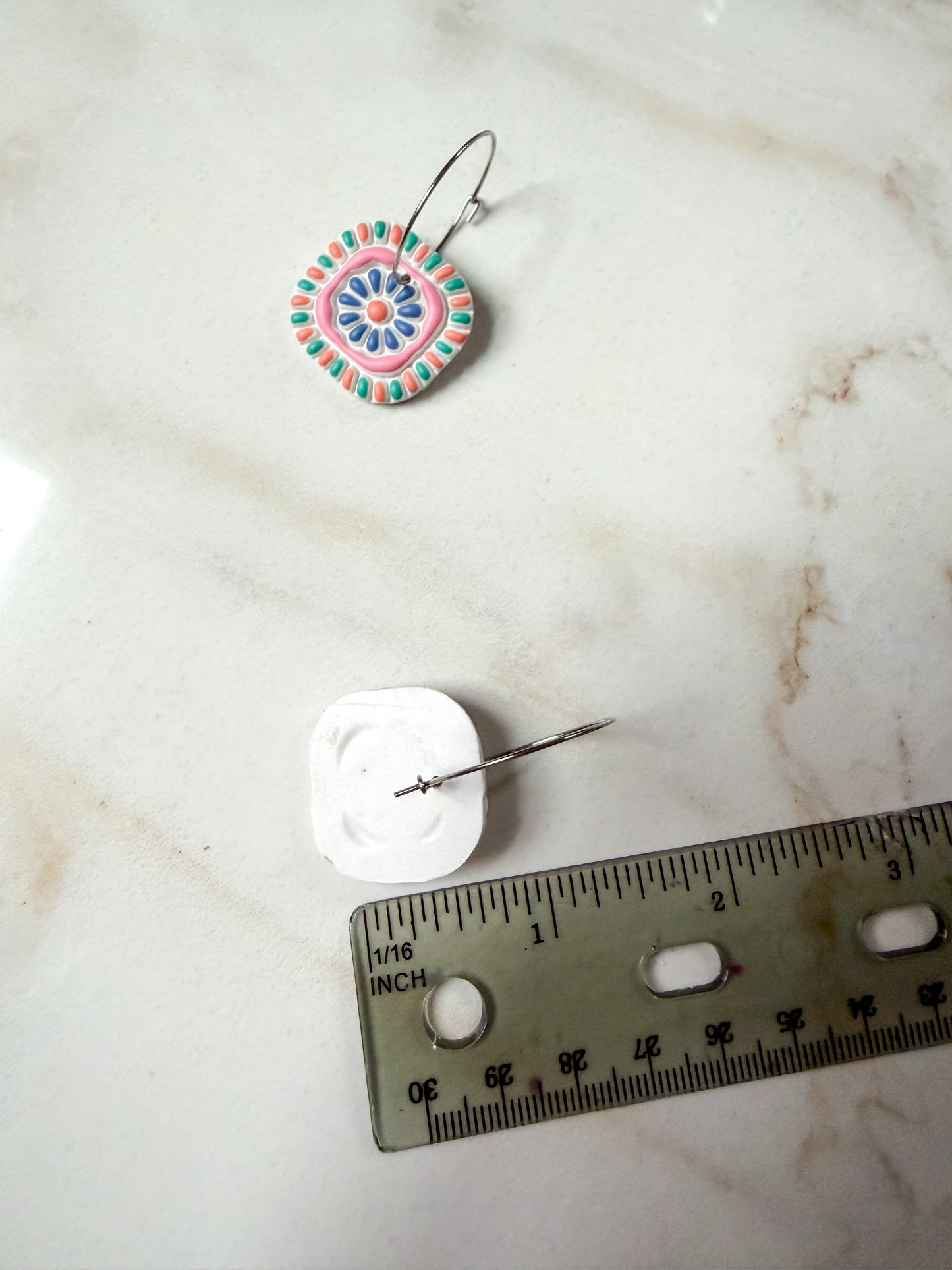 Colorful polymer clay earring on a marble surface with a ruler for scale. About an inch and a half long.