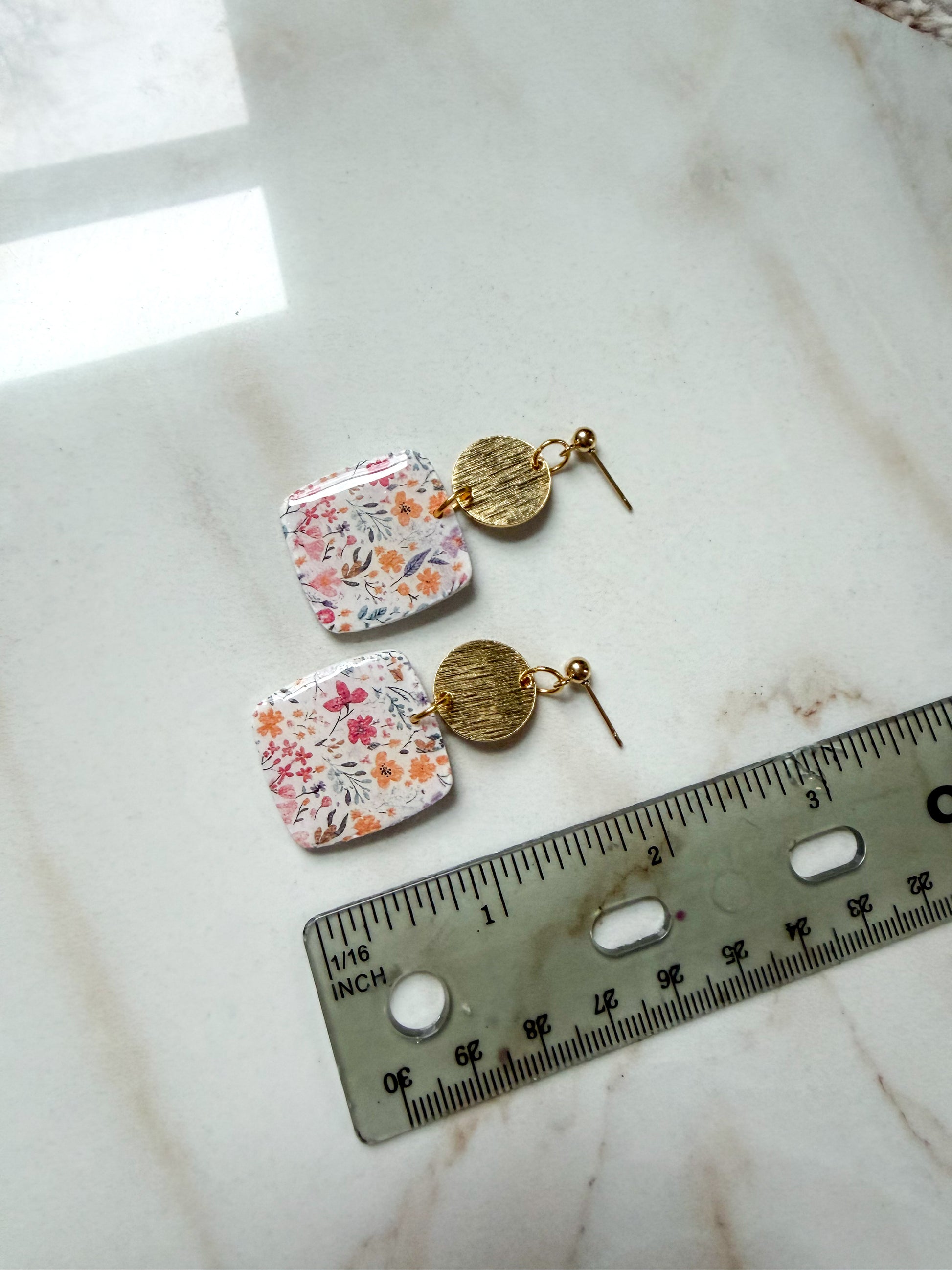 Pair of floral earrings with gold accents on a marble surface next to a ruler for scale.