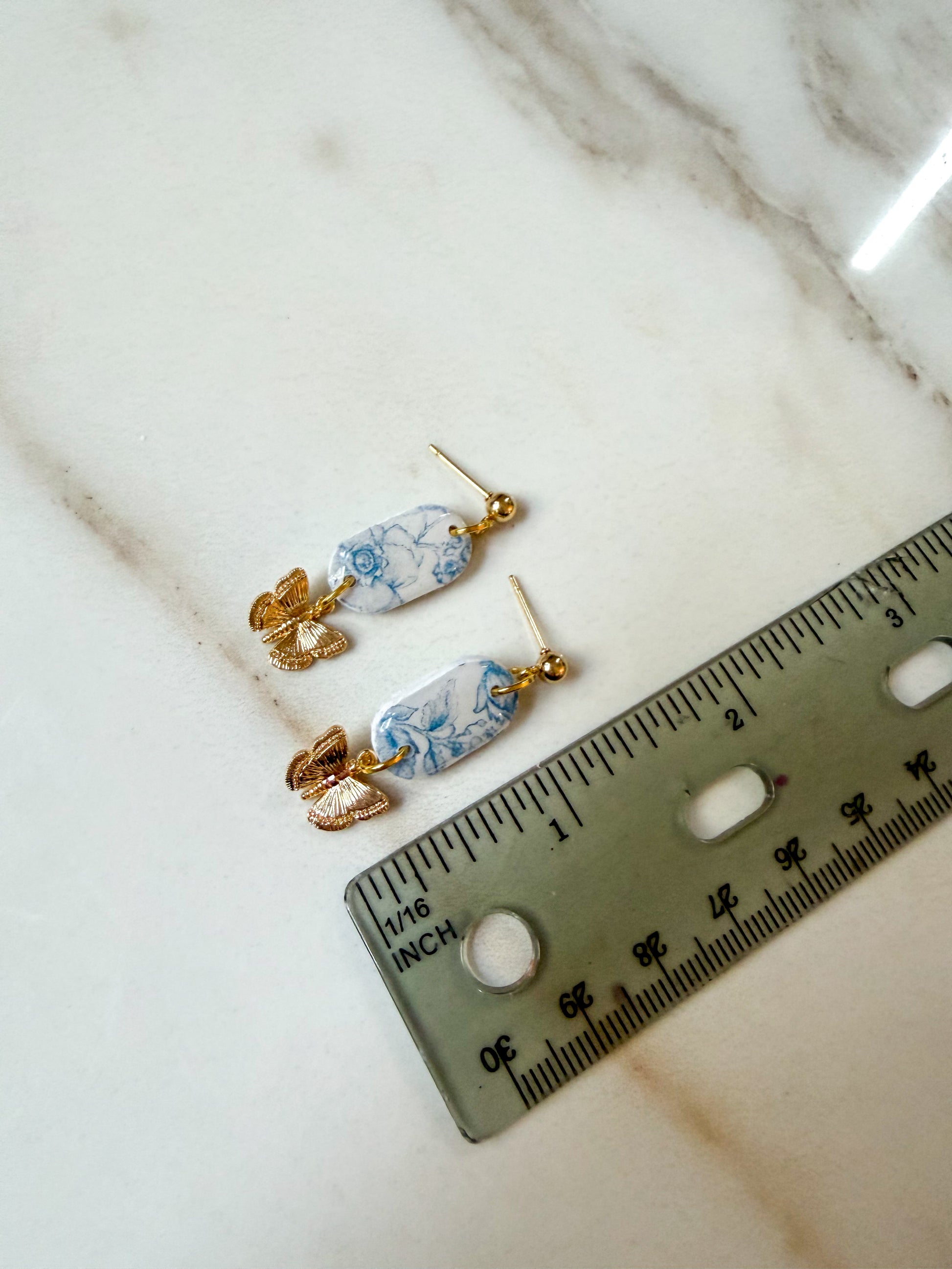 A pair of gold-tone dangle earrings with oval blue-and-white patterned stone pendants, placed on a light marble surface next to a ruler for scale. An inch and a half in length. 