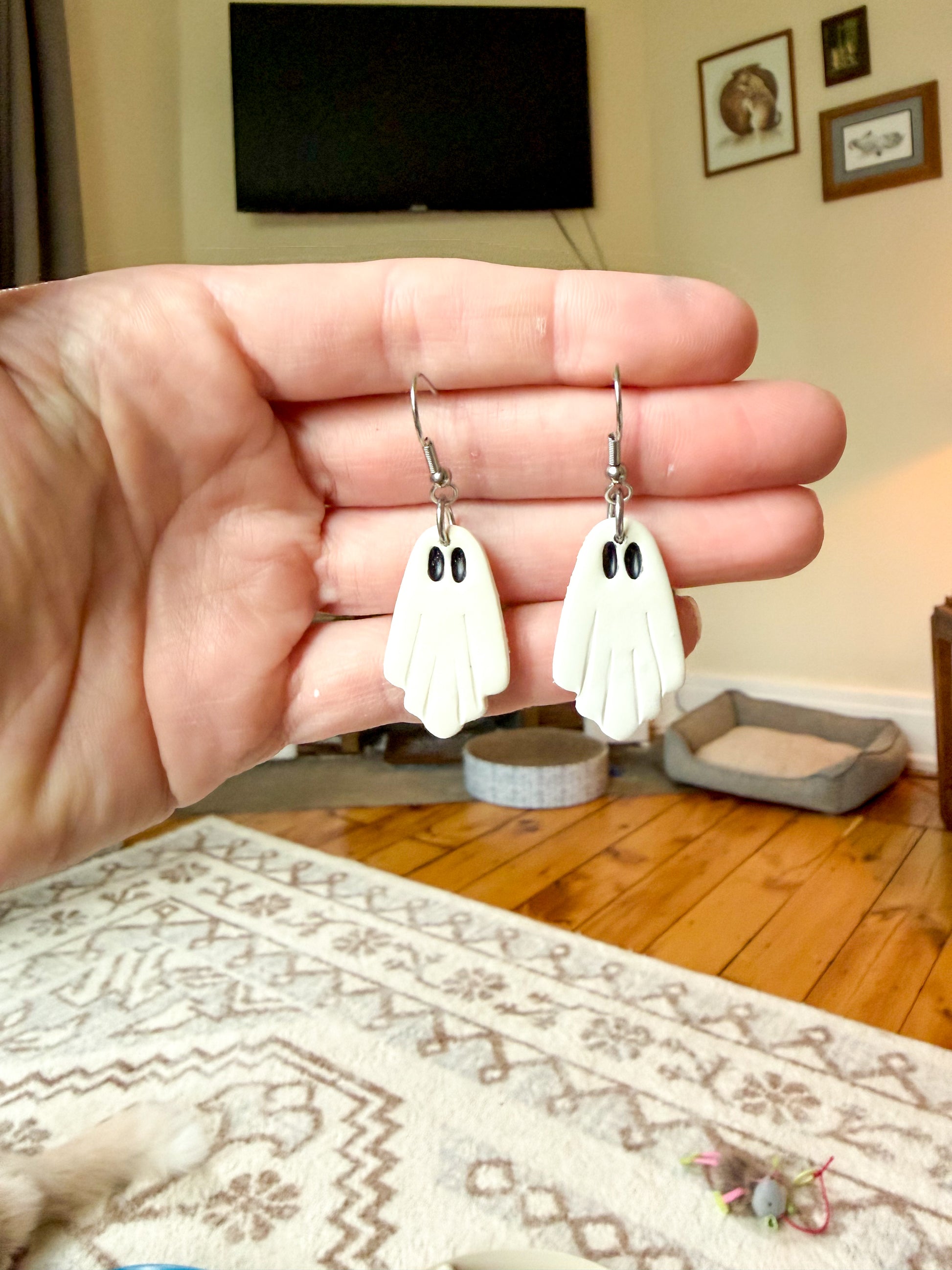 A hand holds a pair of white ghost-shaped dangle earrings with black eyes and silver hooks, shown against a blurred living room background with a TV, rug, and wooden floor.
