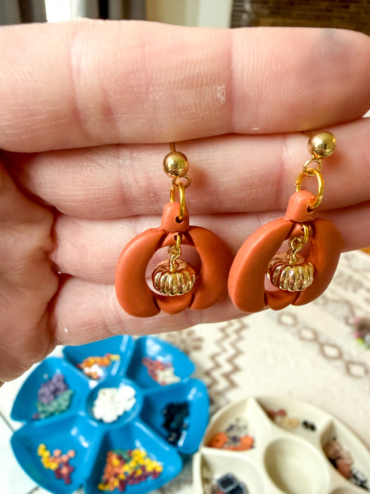 A close-up of a hand holding a pair of orange and gold dangling earrings with small pumpkin charms.