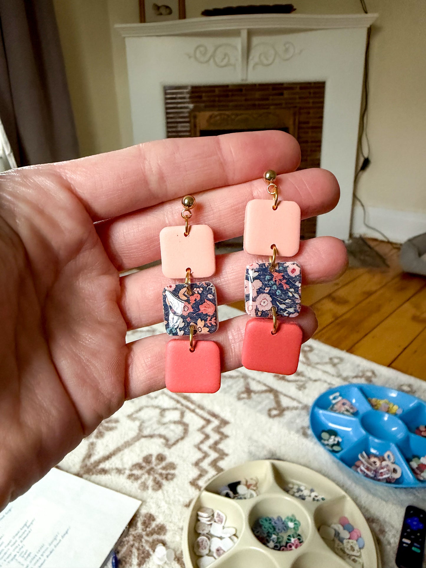Hand holding a pair of colorful square-shaped earrings with a fireplace in the background.