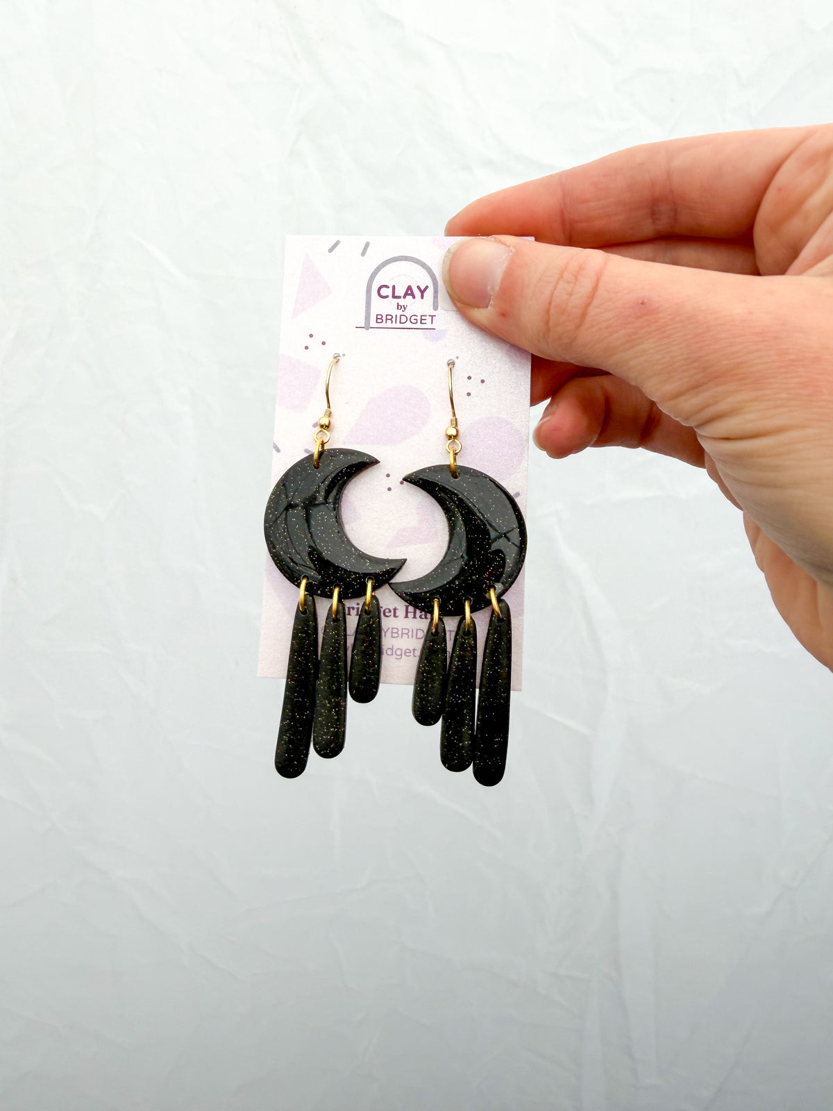 Moon shaped dangle earrings made of sparkly black clay. 3 long pieces hang from each moon like drops. Held in a hand against a white background. 