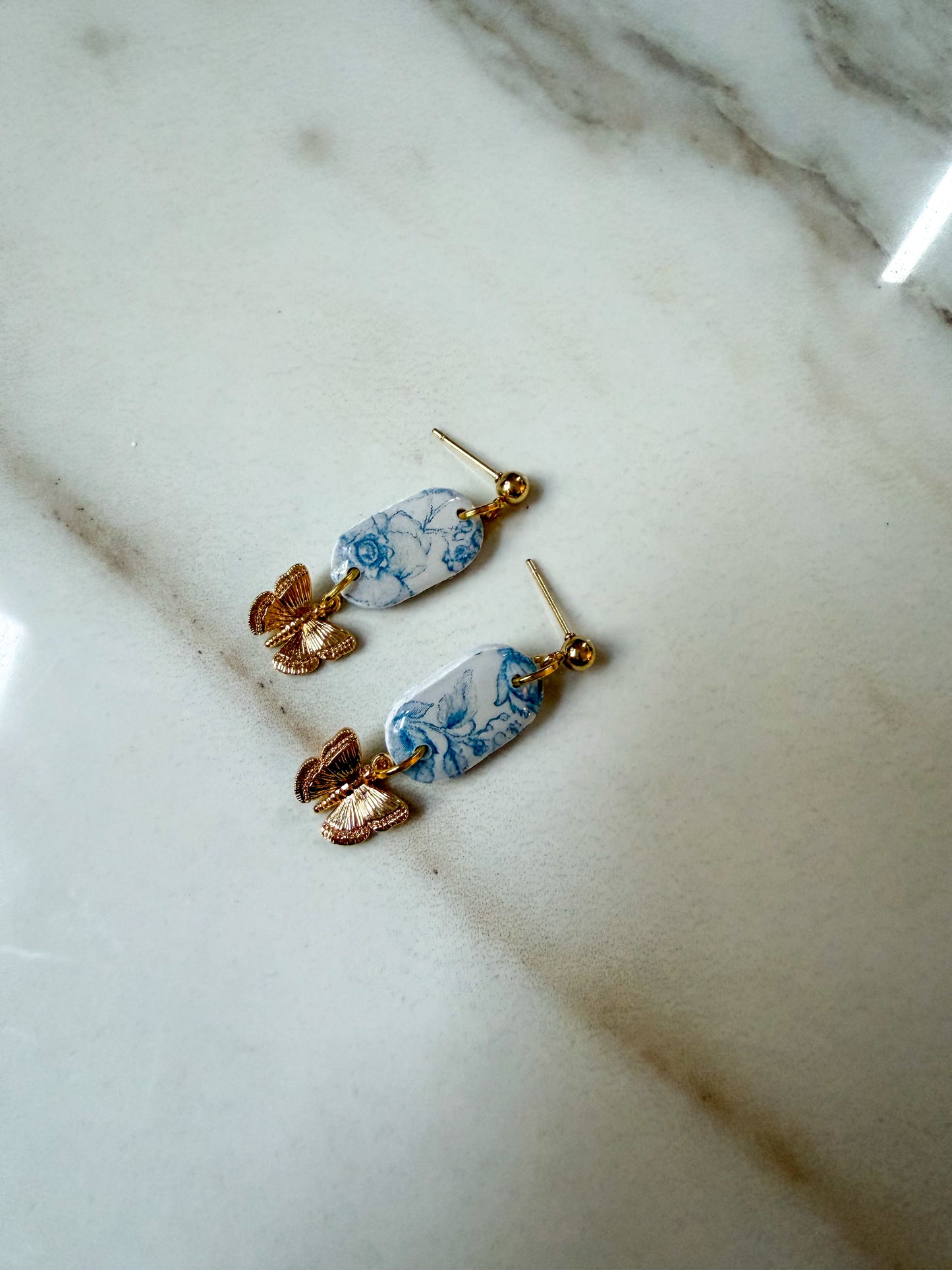 A pair of dangling earrings with blue and white marbled oval charms, gold-tone posts, and rose gold butterfly accents, resting on a white marble surface.