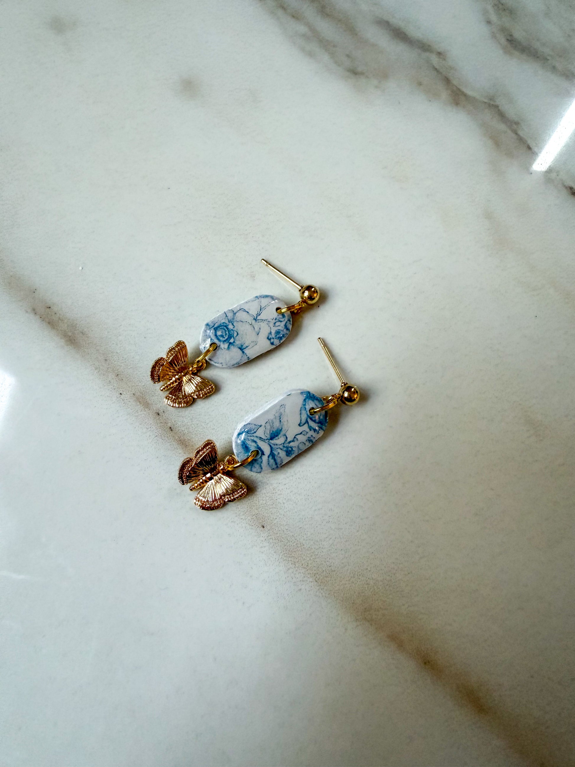 A pair of dangling earrings with blue and white marbled oval charms, gold-tone posts, and rose gold butterfly accents, resting on a white marble surface.