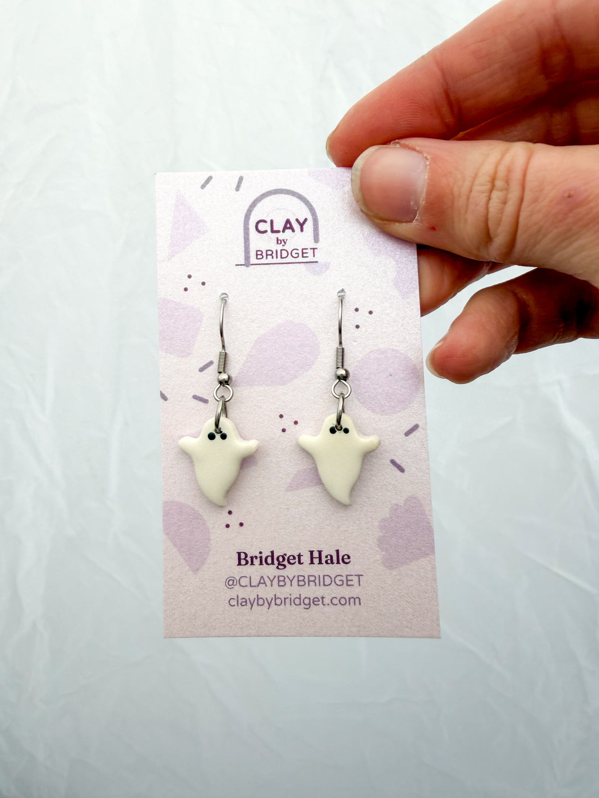 Hand holding an earring card with two white ghosts on silver fishhooks. 
