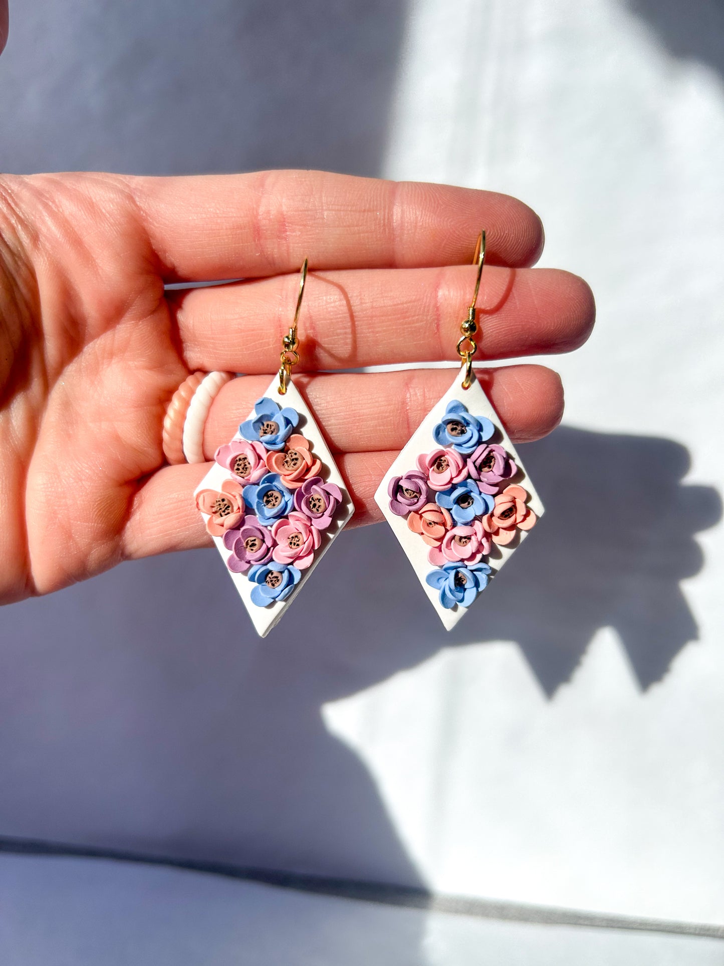 Hand holding a pair of diamond-shaped earrings with floral design against a light background