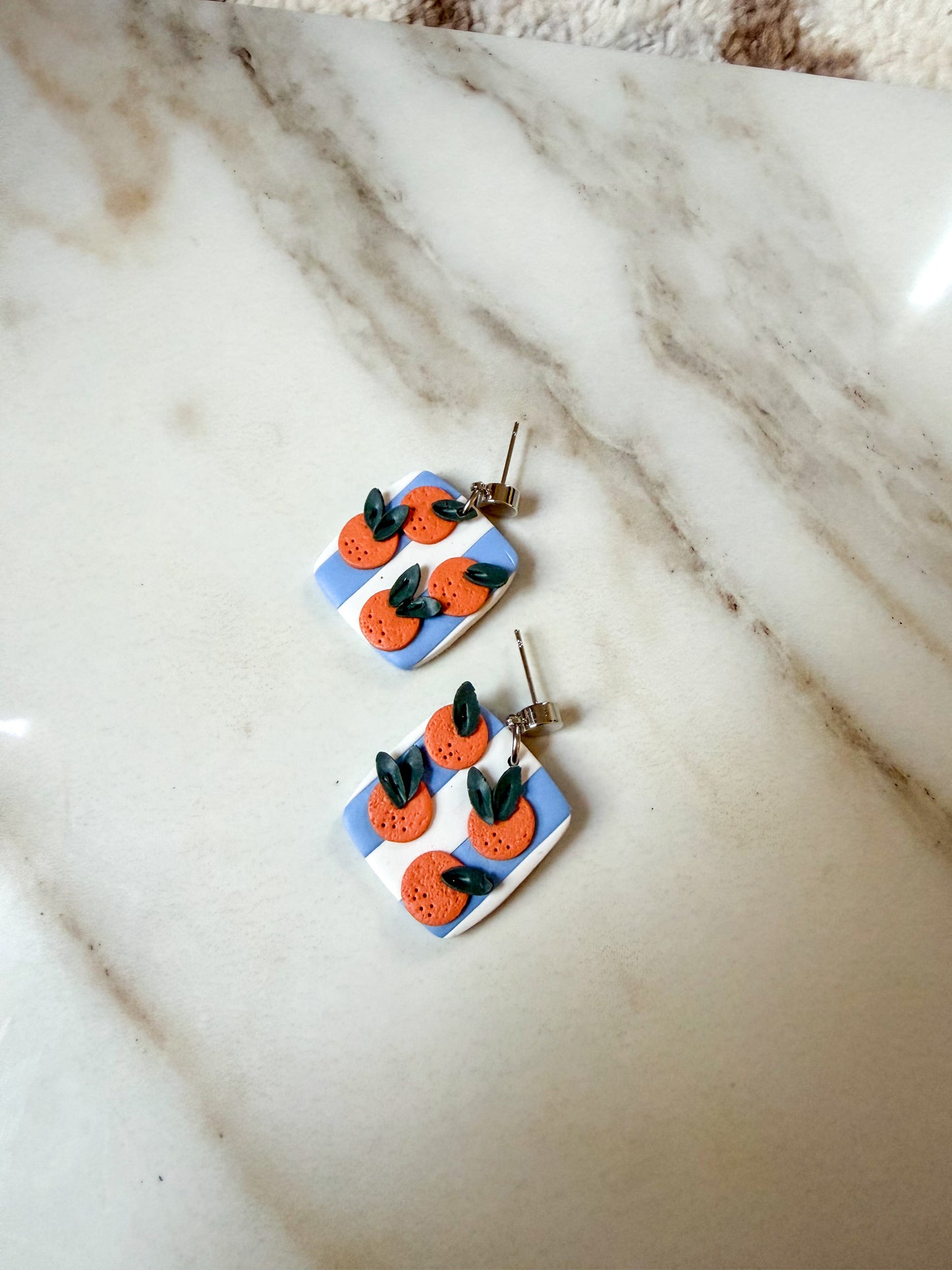 A pair of handmade square earrings featuring a blue and white striped background with orange citrus fruits and green leaves, displayed on a white marble surface.
