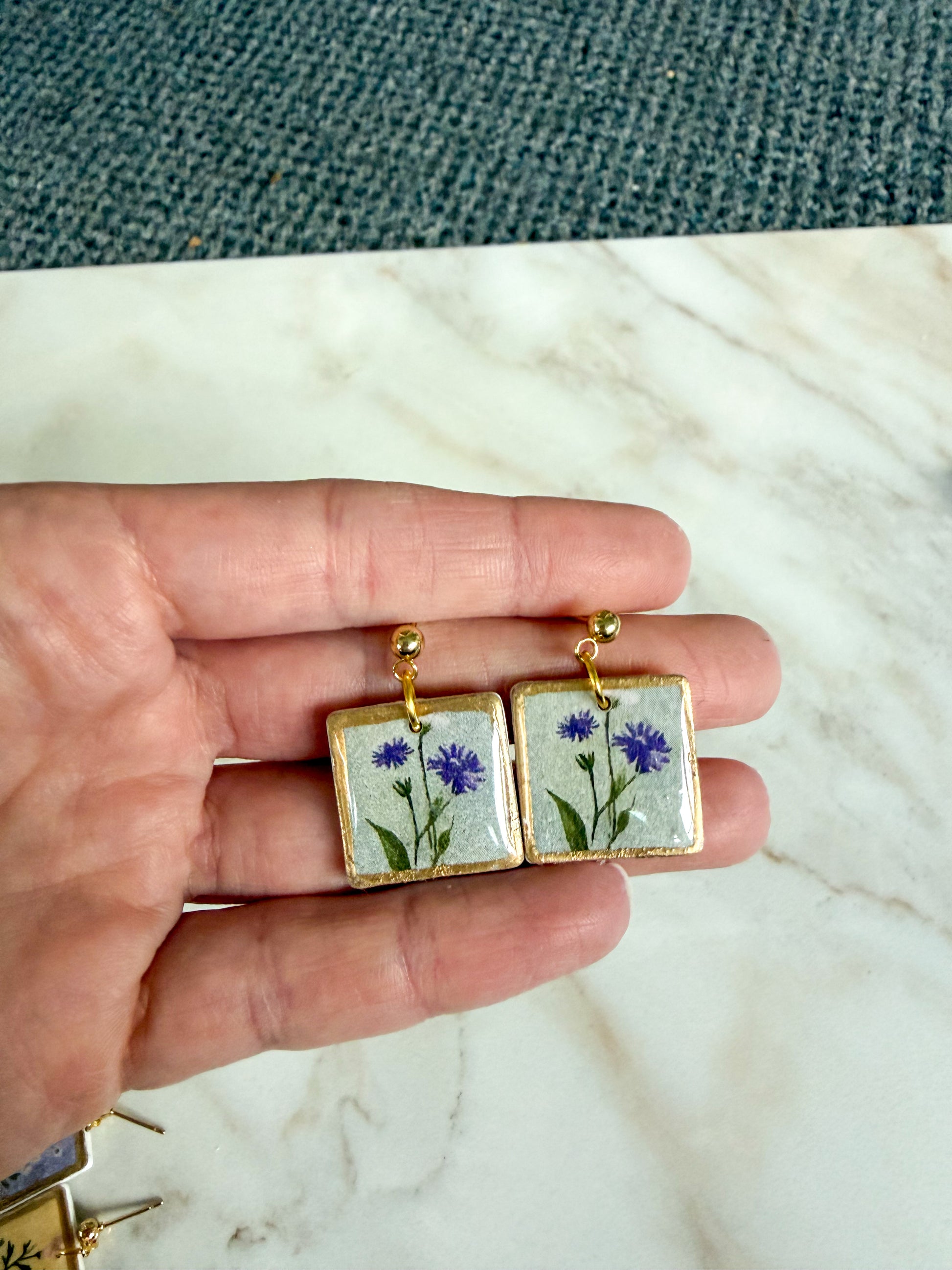 Hand holding a pair of earrings with floral design on a marble surface