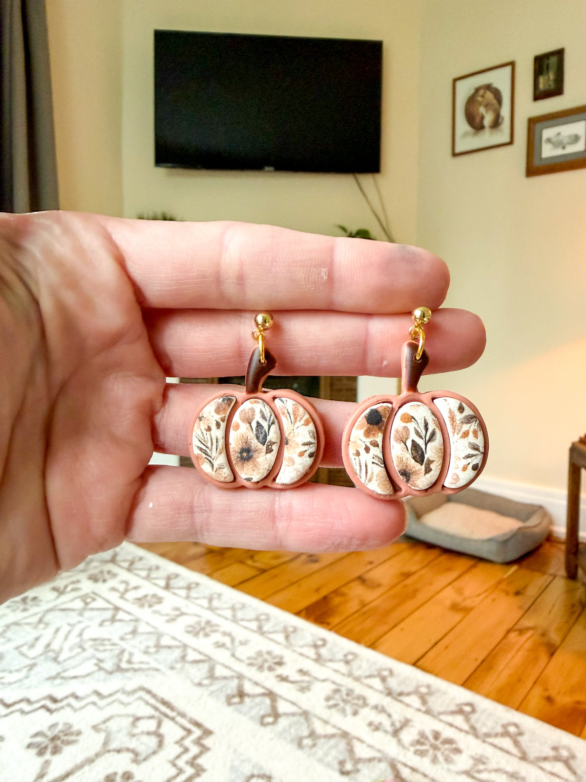 Hand holding pumpkin shaped earrings with floral designs against a living room background. 