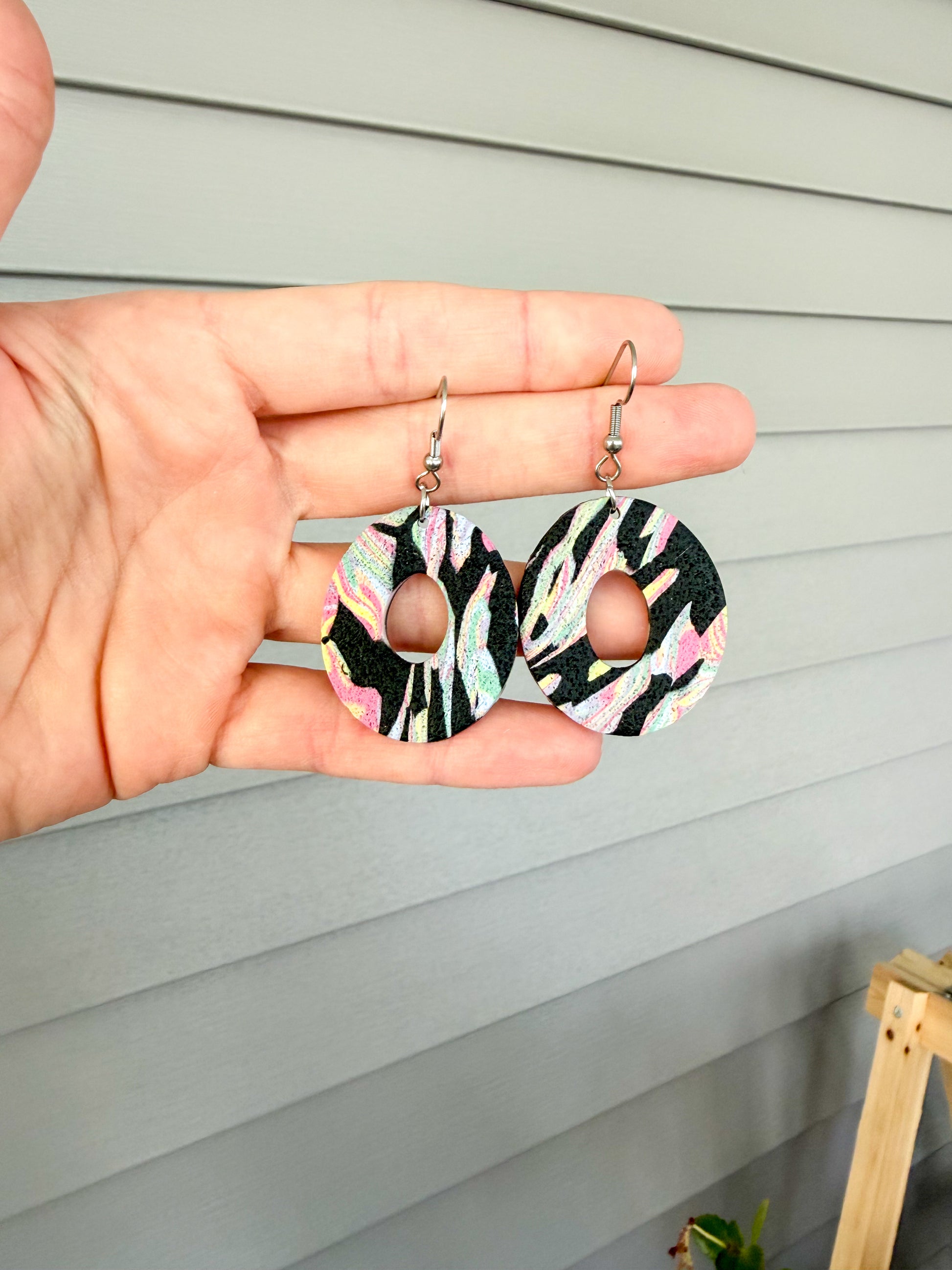 Colorful oval dangles with a center hole cut out. The pattern is black with colorful swirls. They are held in a hand.