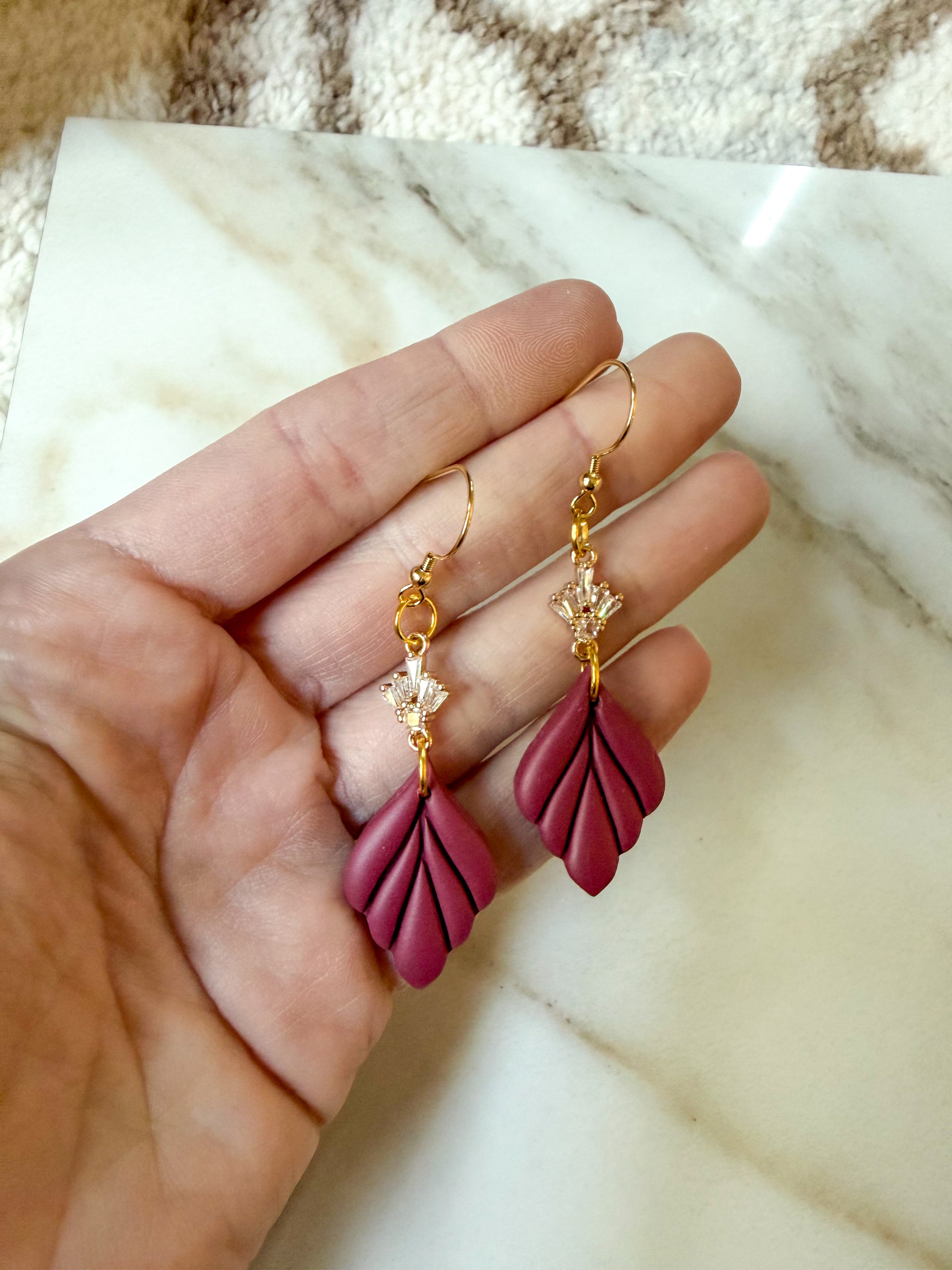 A pair of gold-tone drop earrings with magenta leaf-shaped charms and small clear crystal details, held in a person's hand over a white marble surface.