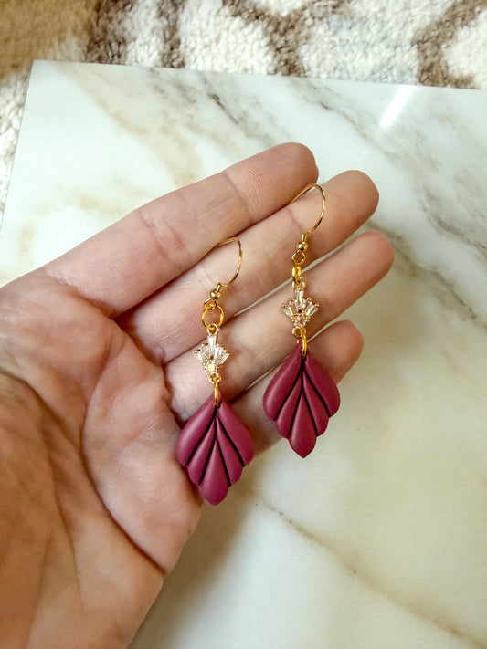 A pair of gold-tone drop earrings with magenta leaf-shaped charms and small clear crystal details, held in a person's hand over a white marble surface.