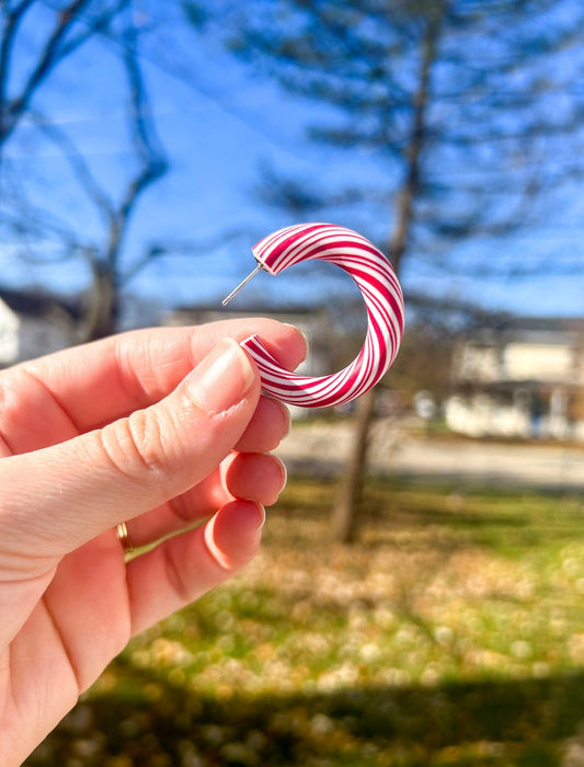 Hand holding a red and white striped earring with a blurred outdoor background. The earrings look like twisted peppermints. 