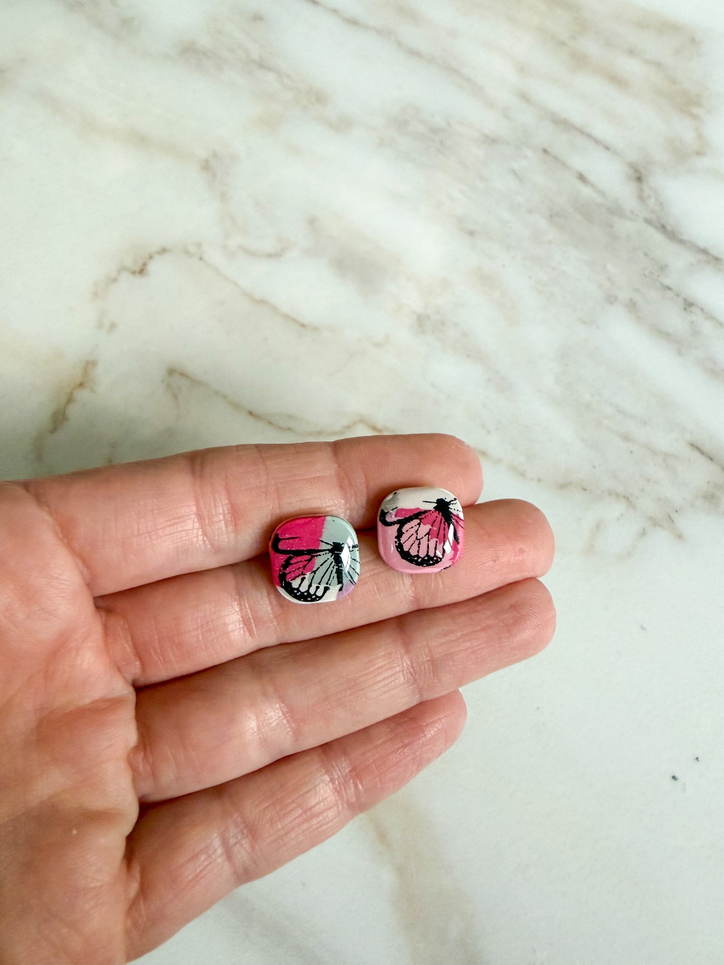 Colorful stud earrings with a butterfly pattern, held in a hand against a marble surface