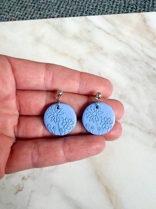 Hand holding two blue circular earrings with floral designs on a marble surface.