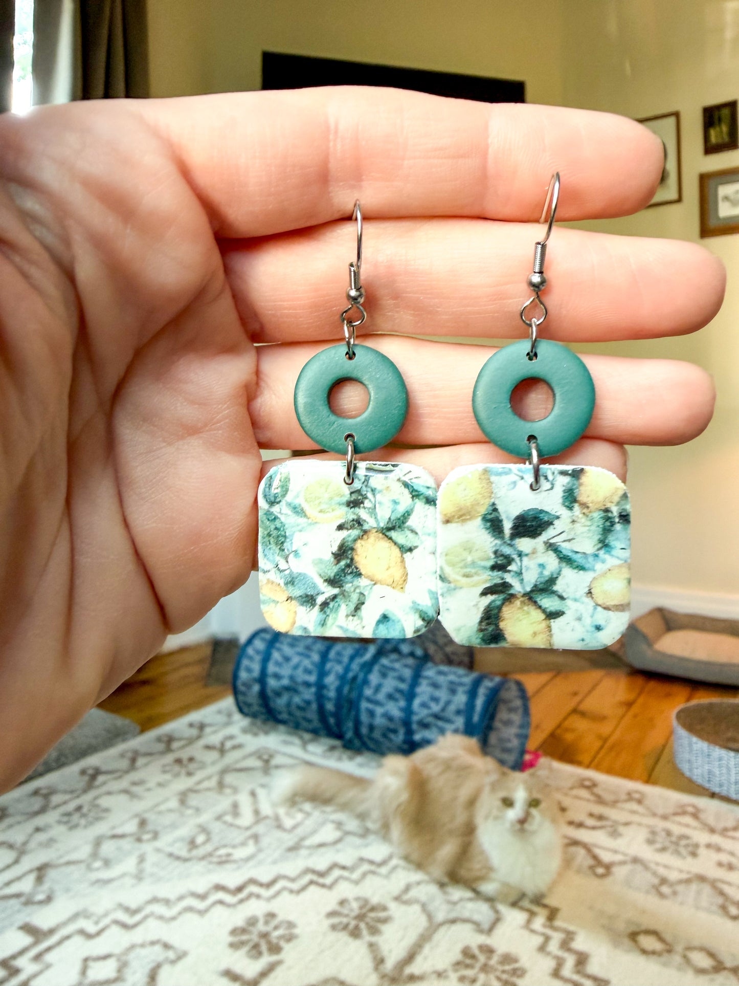 A hand holds a pair of dangling earrings with round turquoise-green top pieces and rectangular floral print lower pieces with a silver fishhook back, shown in a home setting with a cat and cat toys blurred in the background.