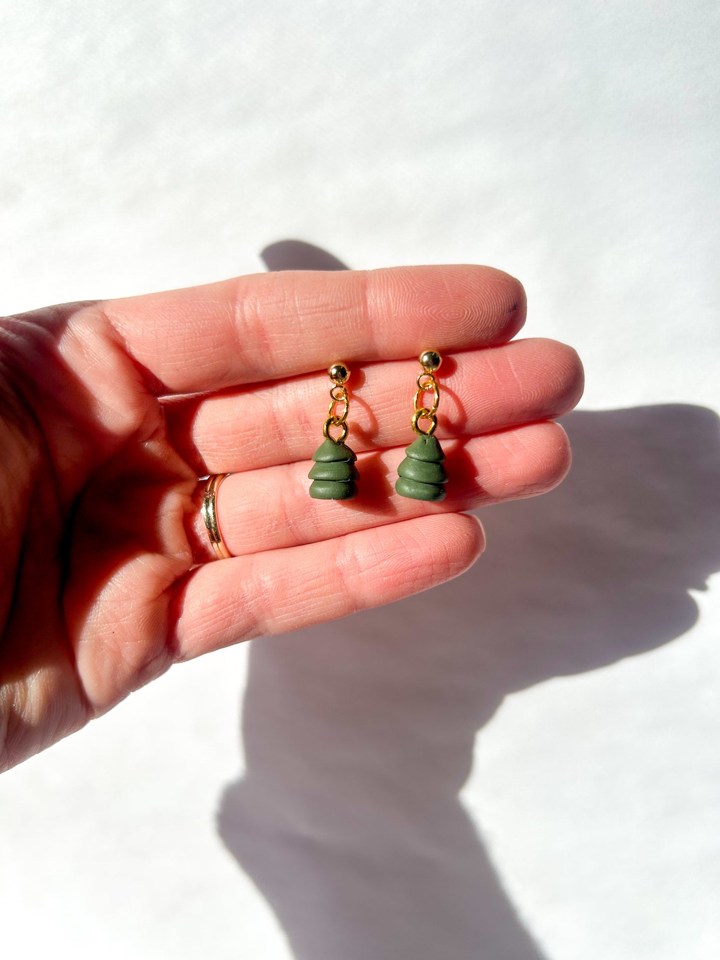 Hand holding two small green earrings with gold accents against a white background. The earrings are small Christmas tree shaped beads.