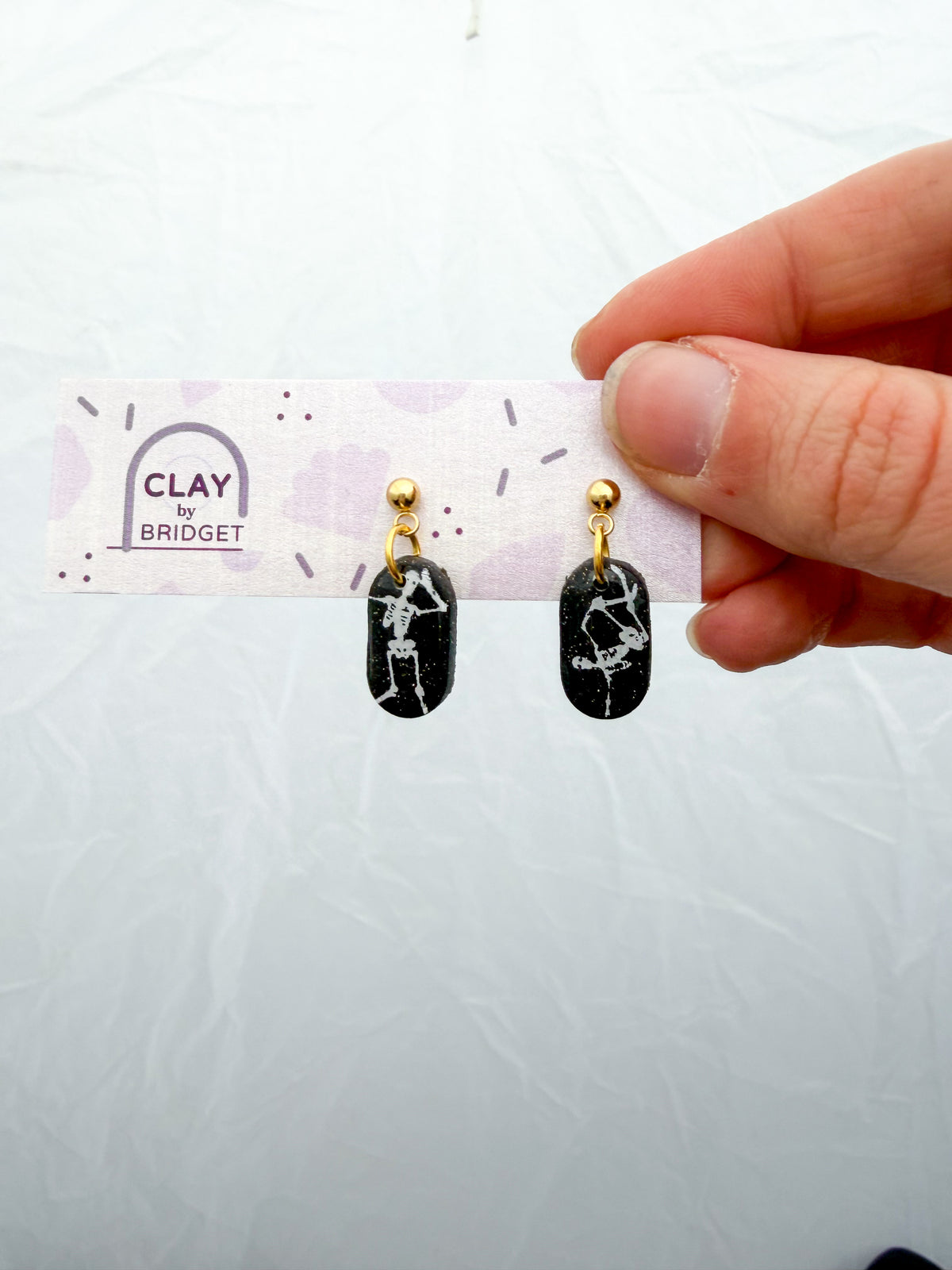 Pill-shaped earrings held in a hand against a white surface. The earrings are glittery black with dancing white skeleton designs on them. 