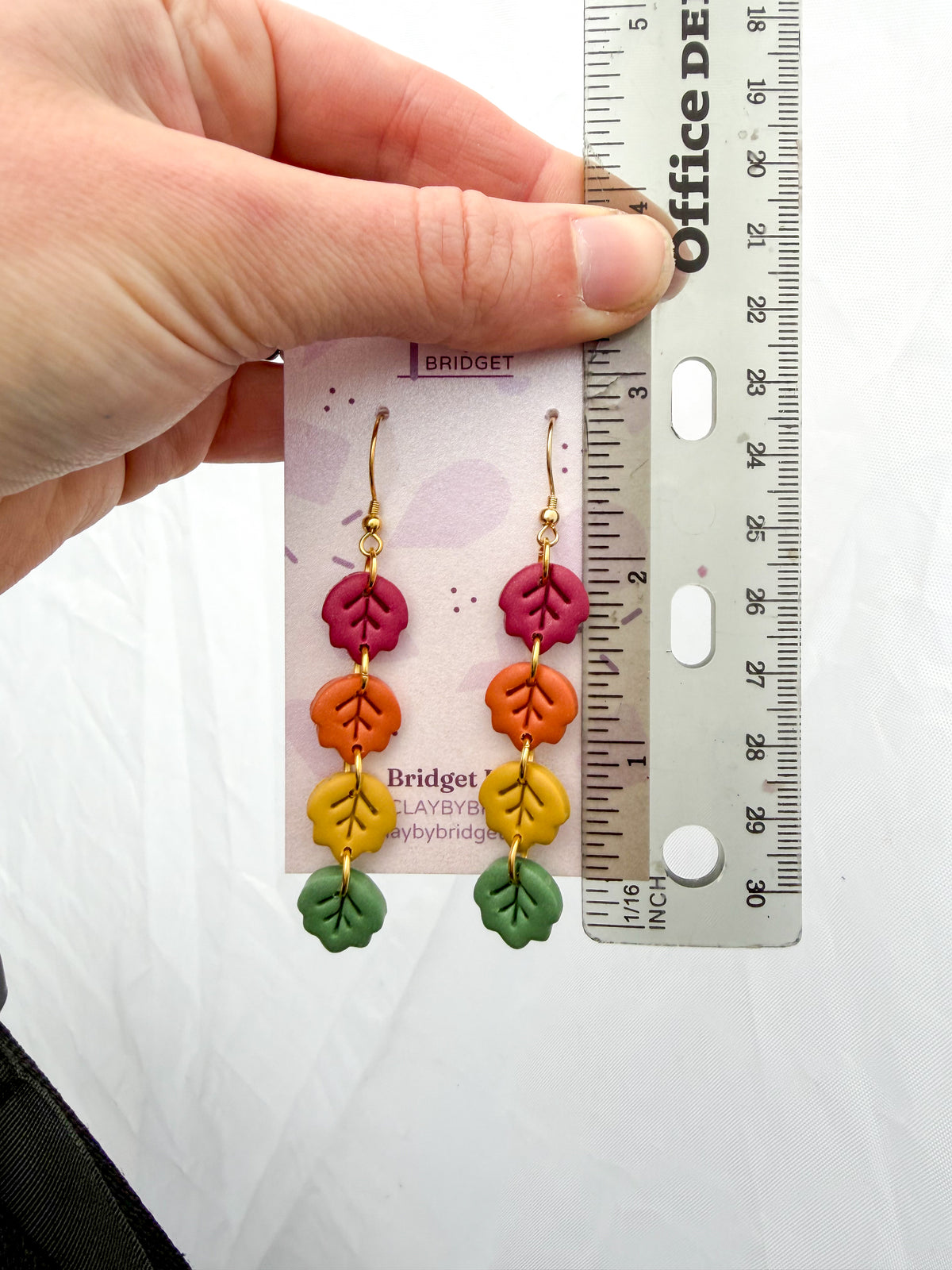 A hand holds a pair of colorful leaf dangle earrings next to a ruler for scale. The earrings feature pink, orange, yellow, and green clay leaves on gold hooks, displayed against a white background. Around 3 inches long. 