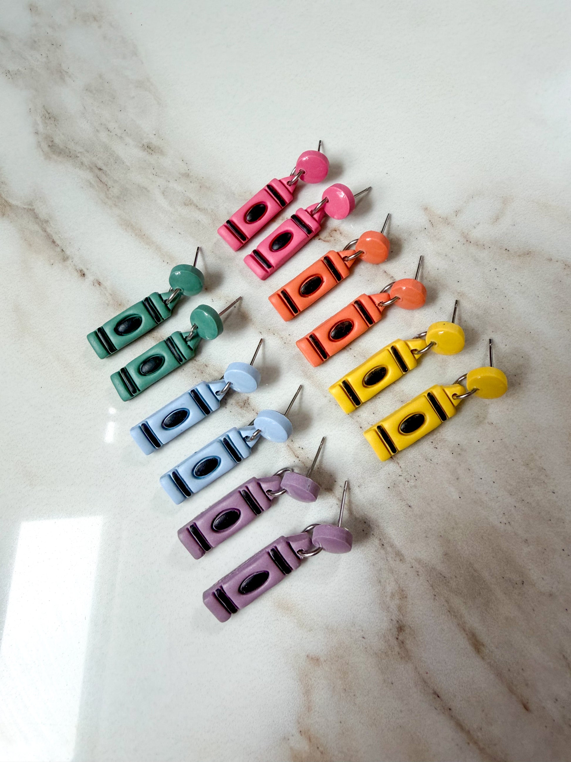 6 pairs of crayon-shaped earrings laying on a marble surface. The colors are pink, orange, yellow, green, blue, and purple. The crayons have a stud top. 