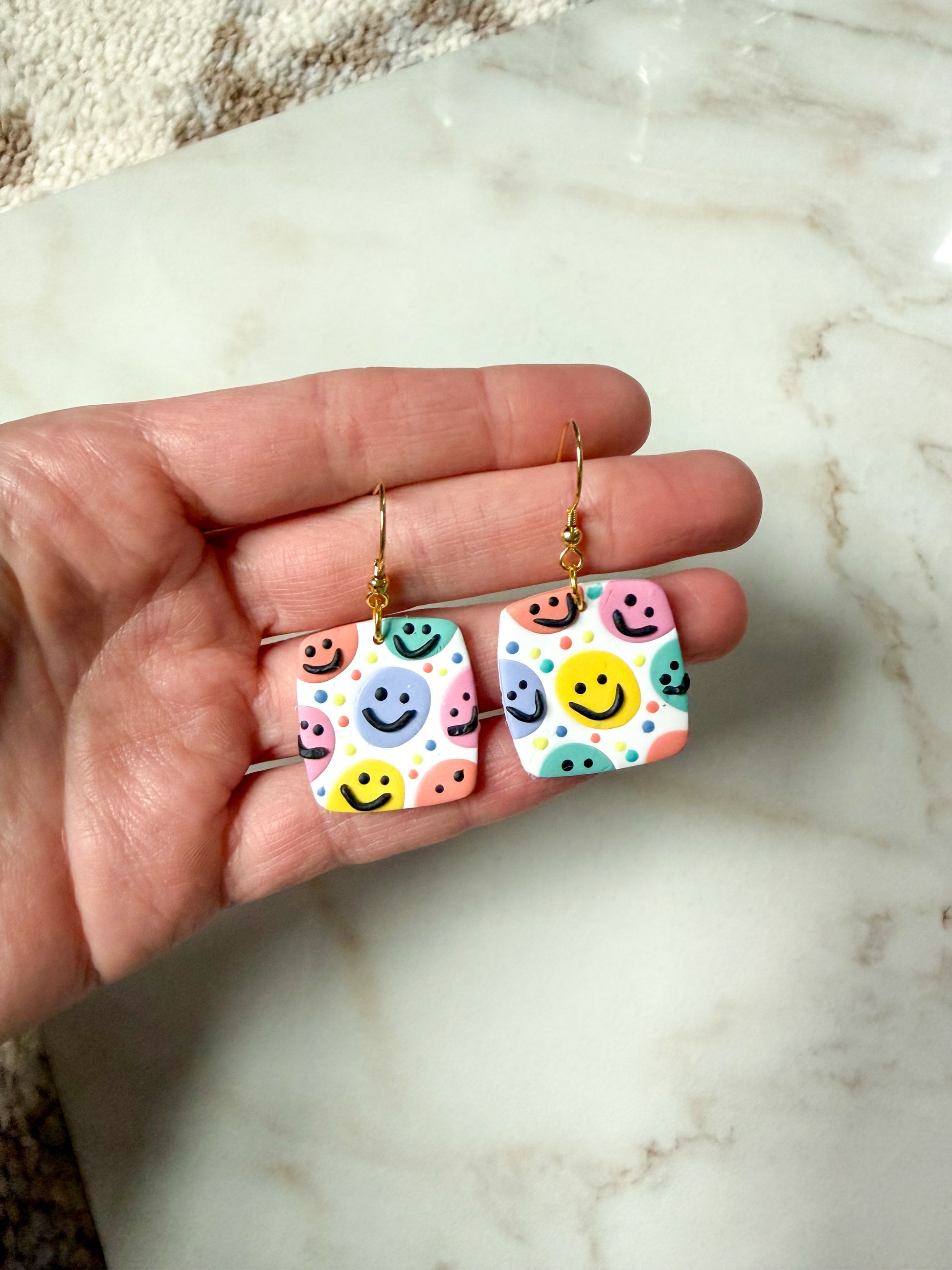 Earrings with a colorful smiley, face design. Held in a hand against a marble surface