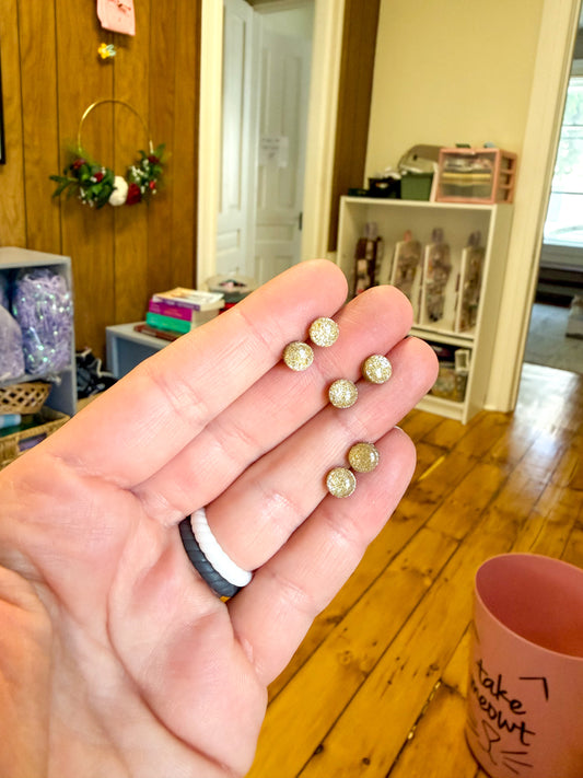 A hand holding six small round stud earrings with textured, glittery gold-toned tops, shown in a home setting with wooden floors.
