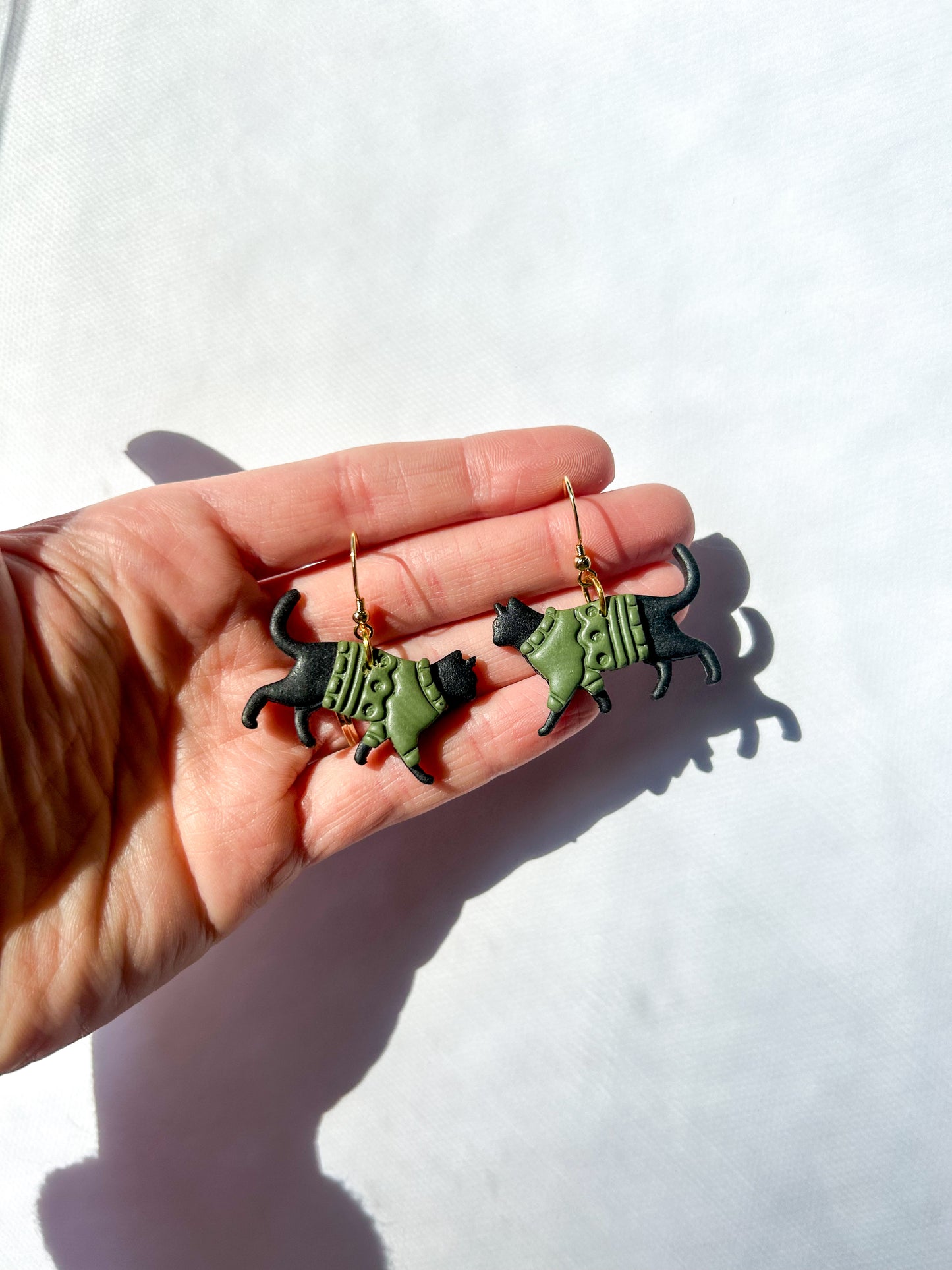 Hand holding two black cat-shaped earrings against a white background. The cats have green sweaters. 