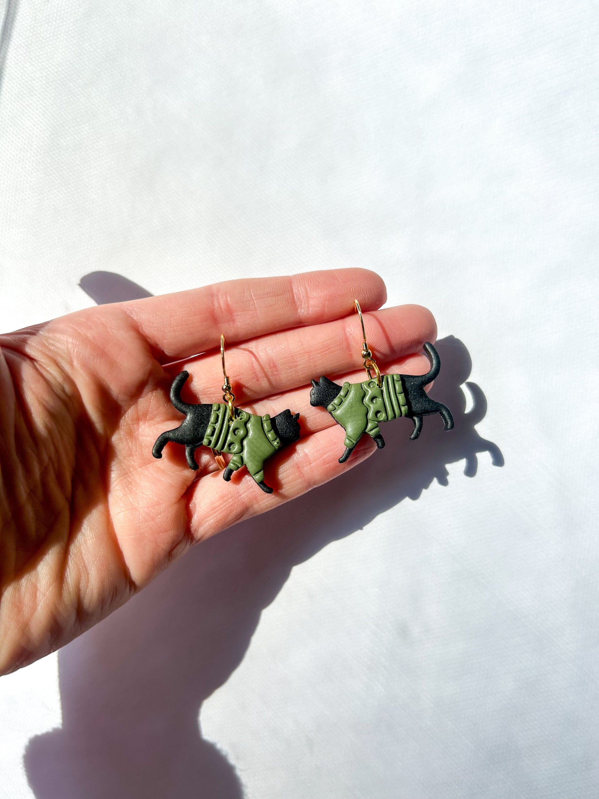 Hand holding two black cat-shaped earrings against a white background. The cats have green sweaters. 