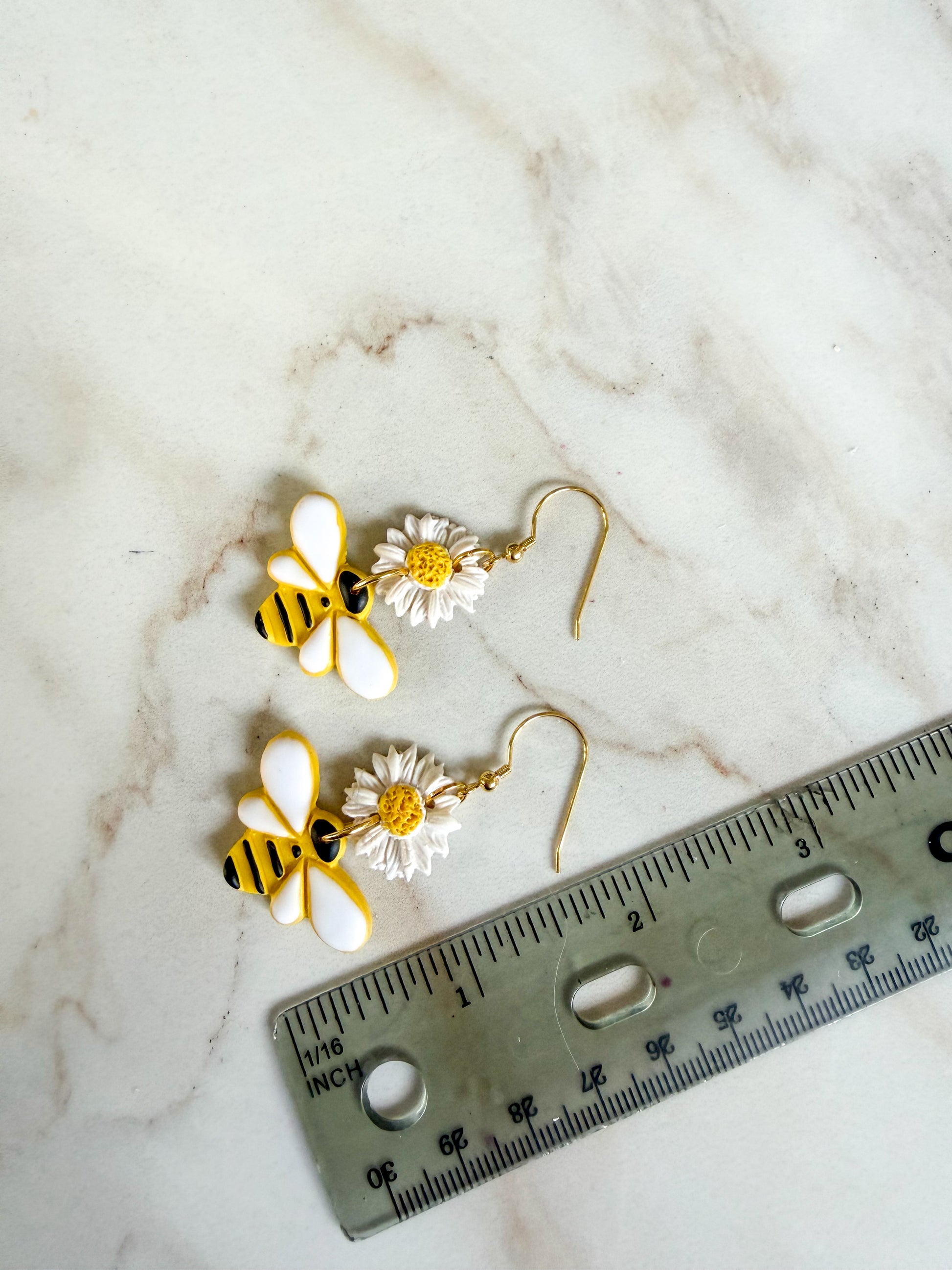 Bee-shaped earrings with flowers on a marble surface next to a ruler for scale. About 2 inches long 