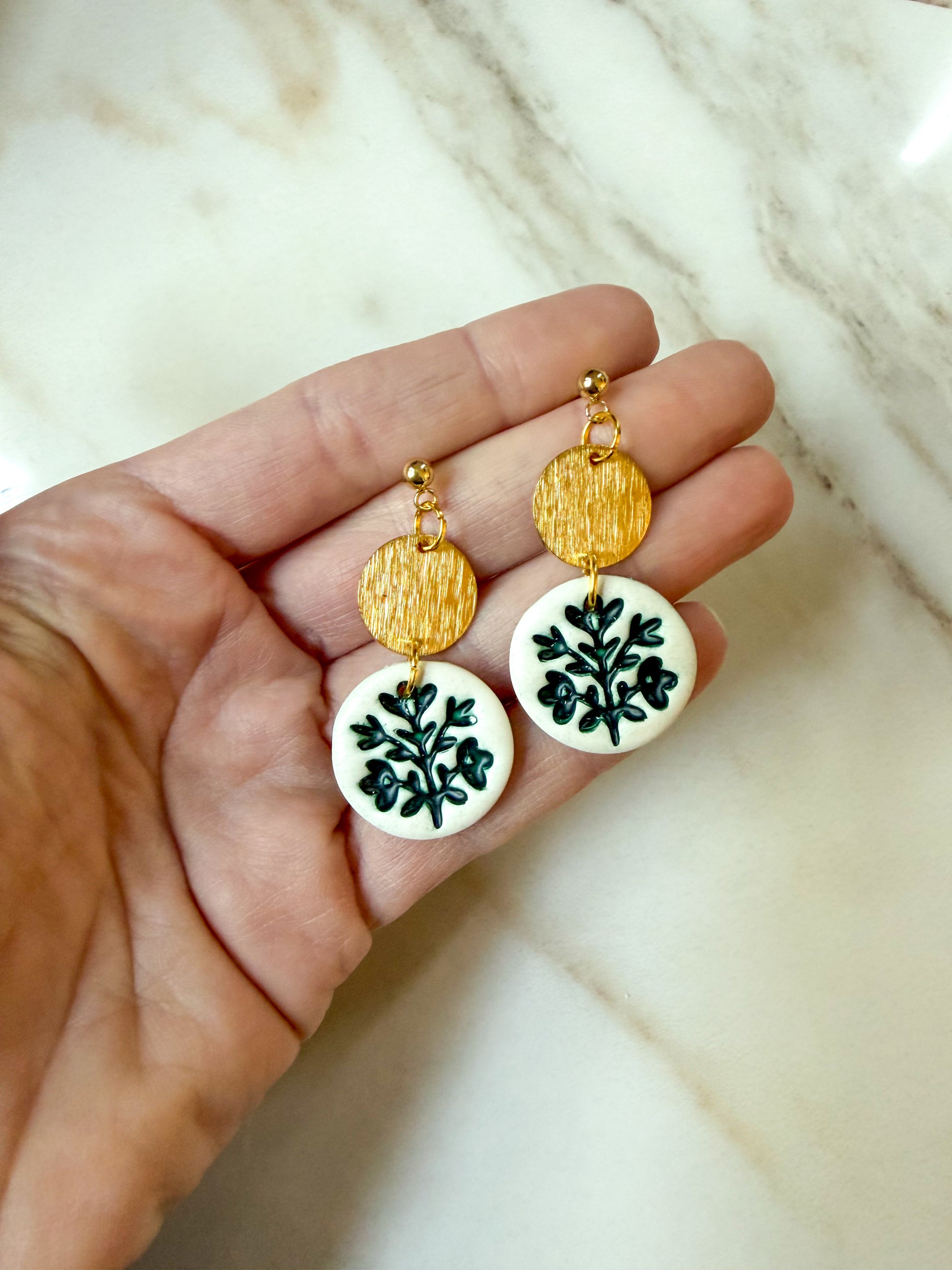 A pair of handmade dangle earrings held in a hand. The earrings feature textured gold circular discs at the top and white circular discs at the bottom, decorated with green leafy branch pattern.