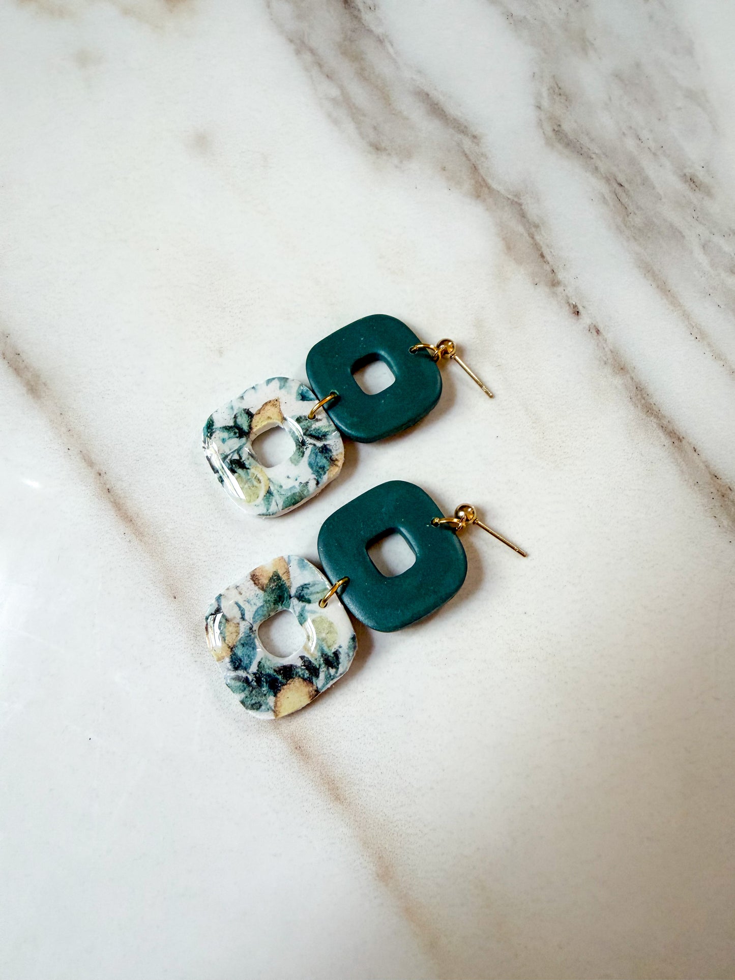 A pair of geometric drop earrings with dark green square charms and white lemon patterned square charms, displayed on a light marble surface.